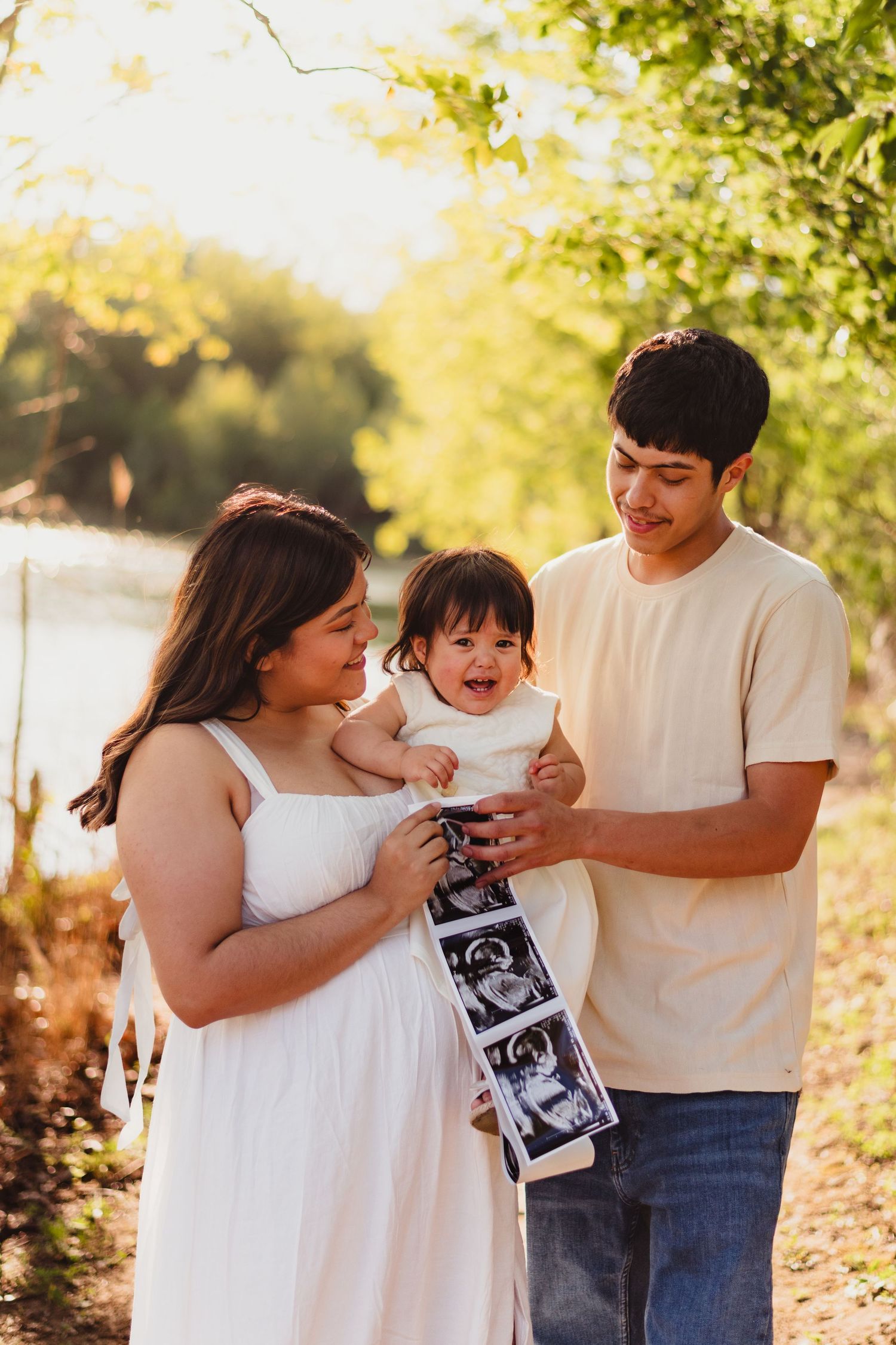 A family shares a joyful moment while holding ultrasound photos outdoors in golden sunlight.
