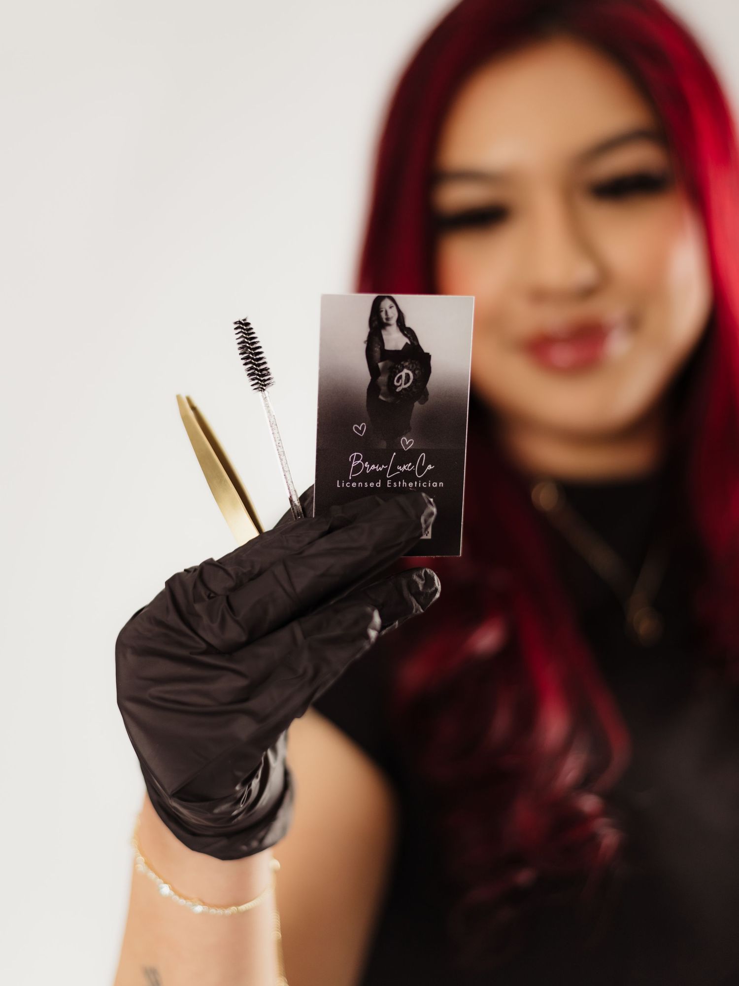 A red-haired esthetician in black gloves holds brow tools and a business card for Brow Lux Co Licensed Esthetician.