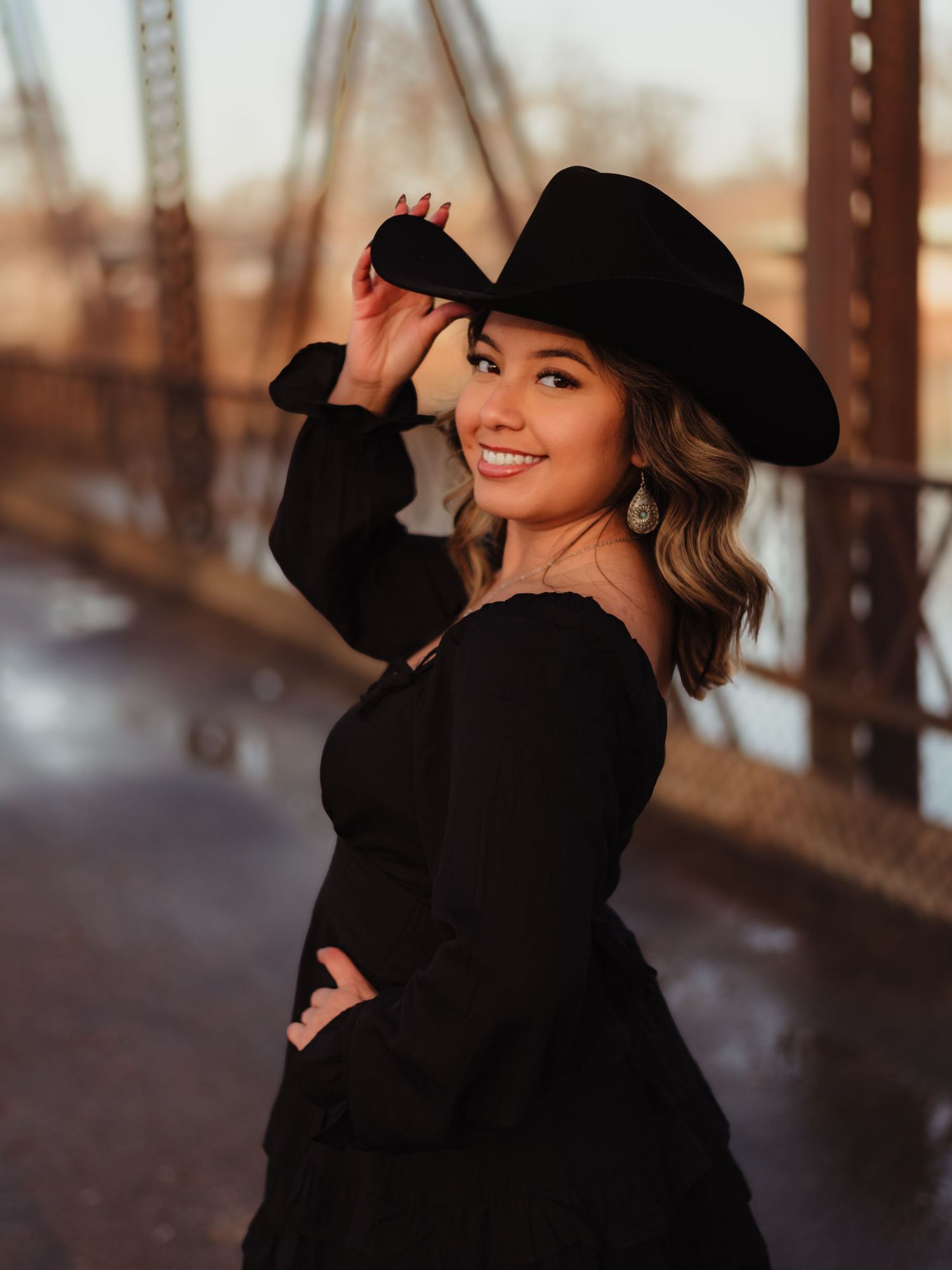 A woman in a black dress and cowboy hat poses on a bridge during golden hour with a warm autumn background.