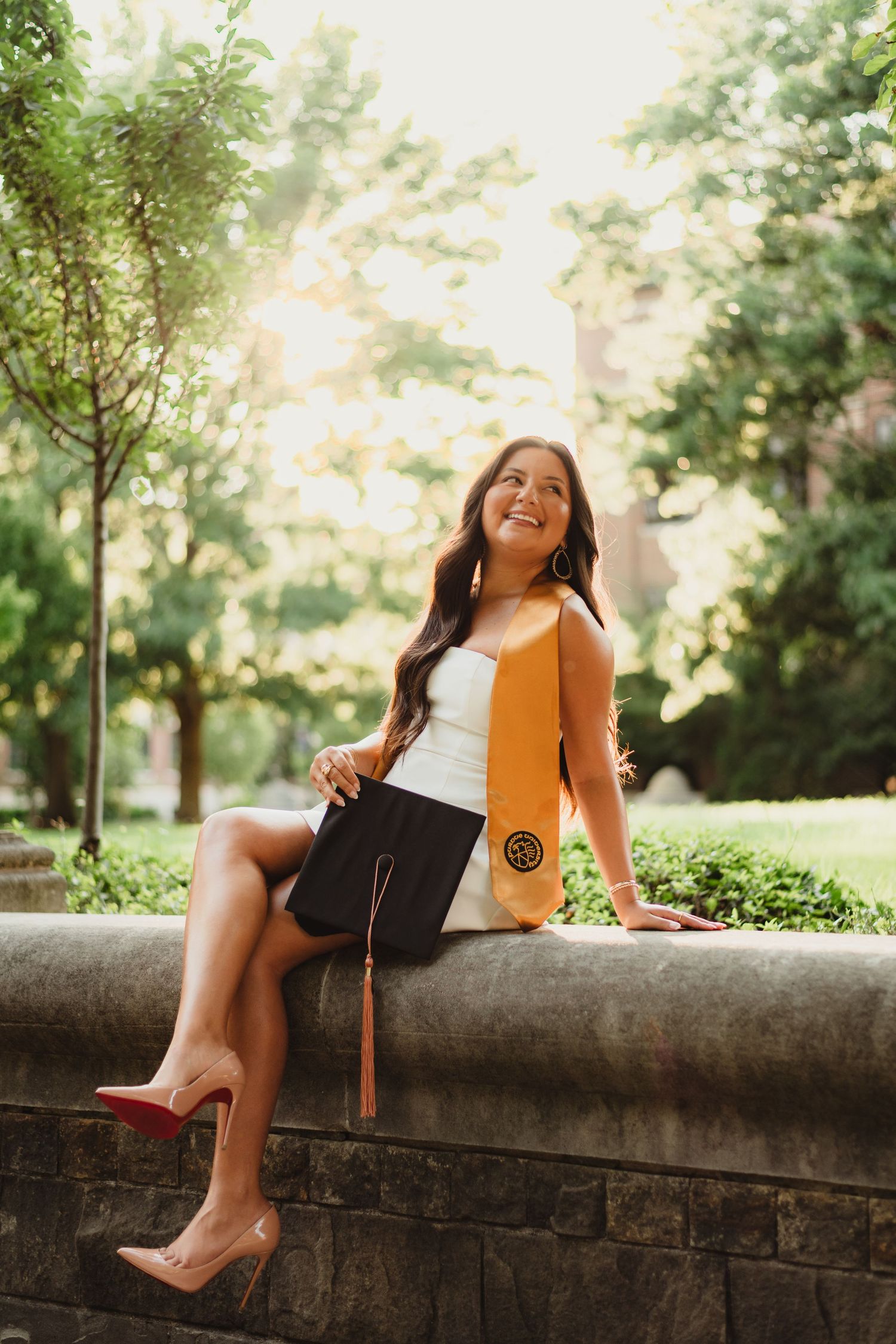 A graduate in cap and gown sits on a stone wall in a sunlit garden setting wearing white top and high heels  at Purdue Campus.