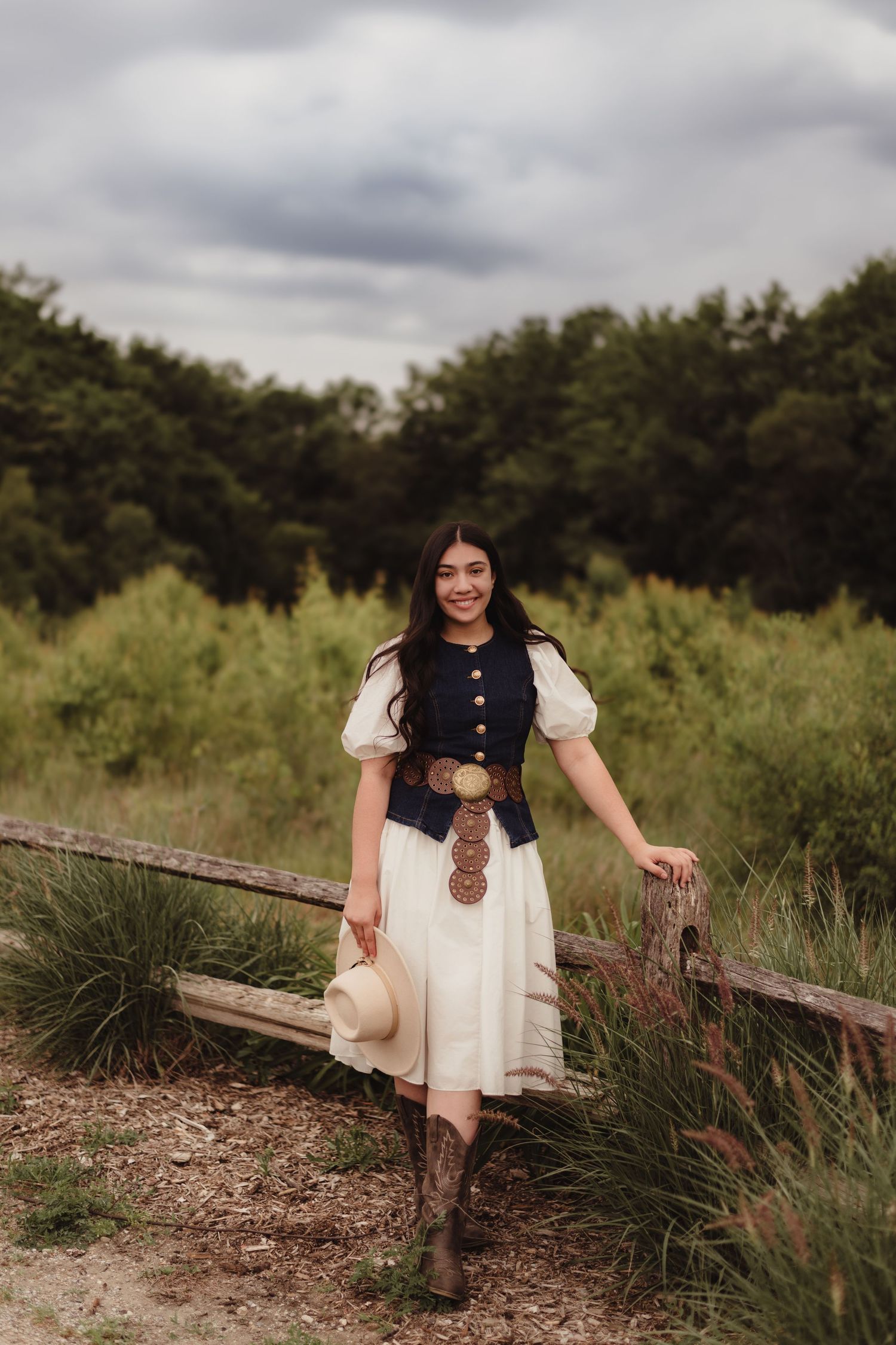 western senior outfit with cowboy boots, white dress, navy vest, and hat poses by rustic fence in green field in Lafayette, indiana