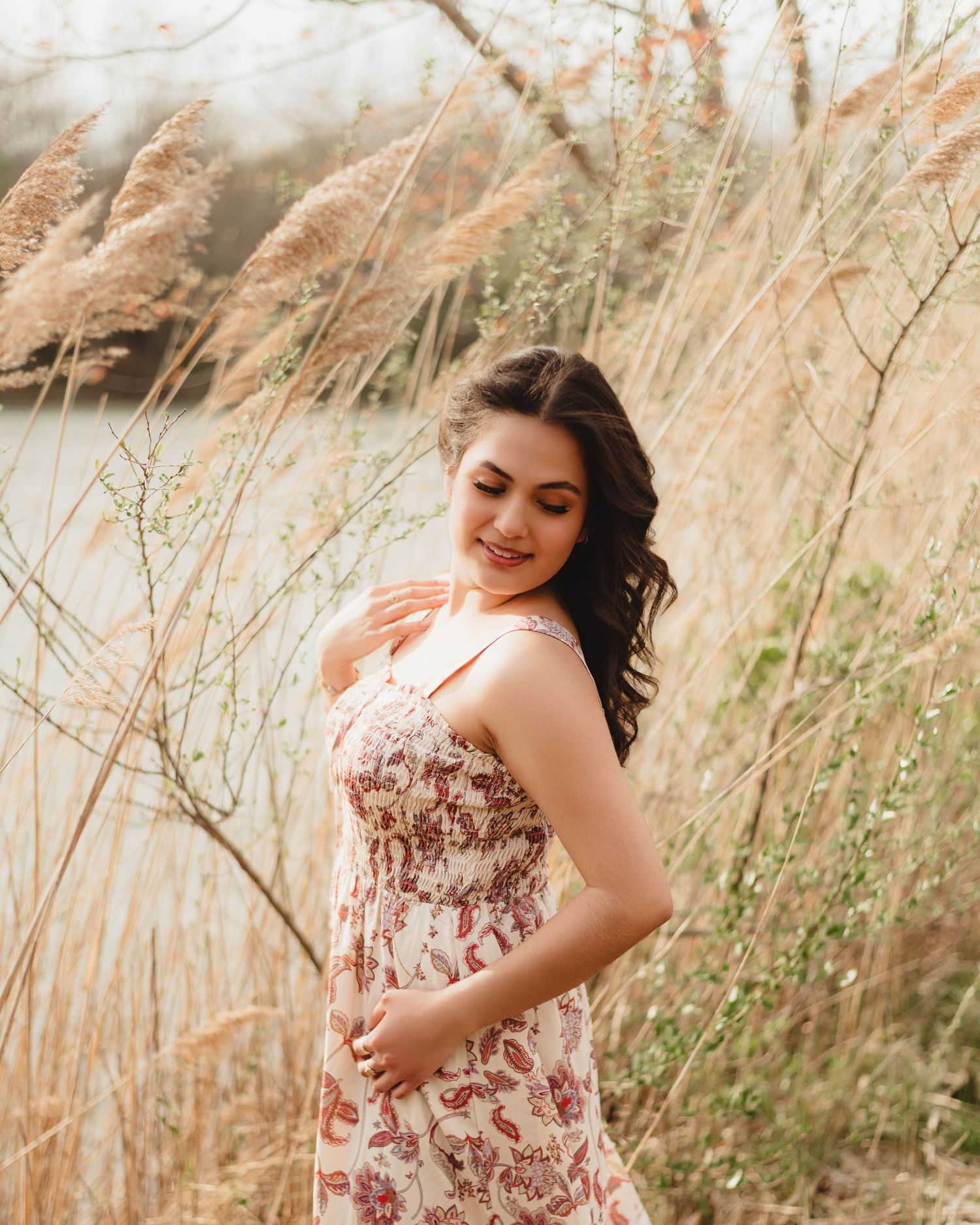 High school senior in floral sundress smiling among tall golden grasses near a serene lake in Lafayette, Indiana