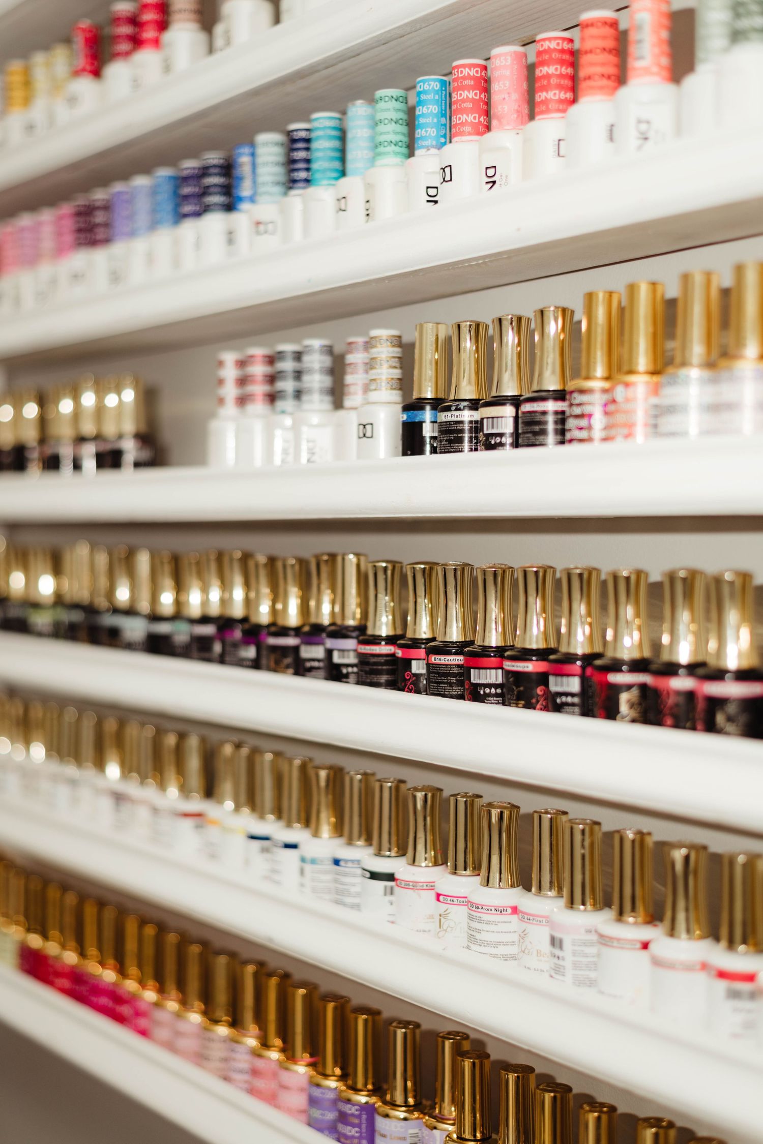 White shelves displaying rows of colorful nail polish bottles with gold caps in a nail salon.