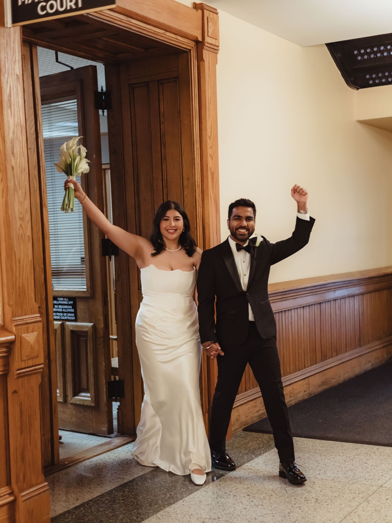 A joyful couple celebrates after their courthouse wedding ceremony, raising their arms in triumph in a hallway.