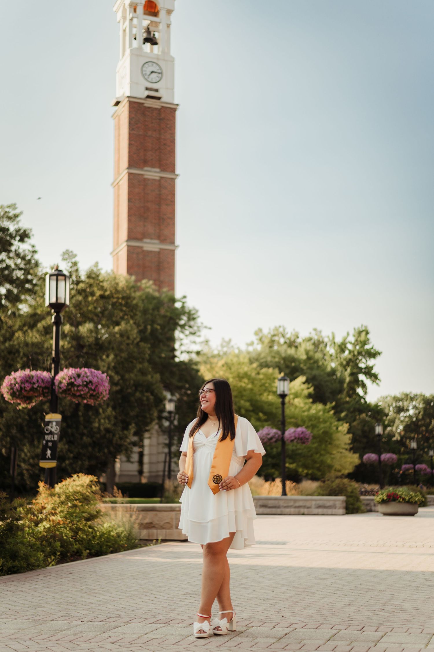 A person in a white dress stands on a brick plaza in front of a Purdue Bell tower with pink flowers on lampposts at Purdue Campus.