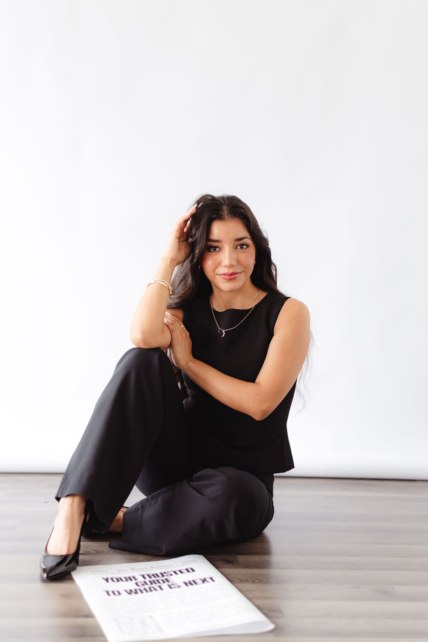 Young woman in black outfit sitting on floor beside newspaper, posing against white wall.