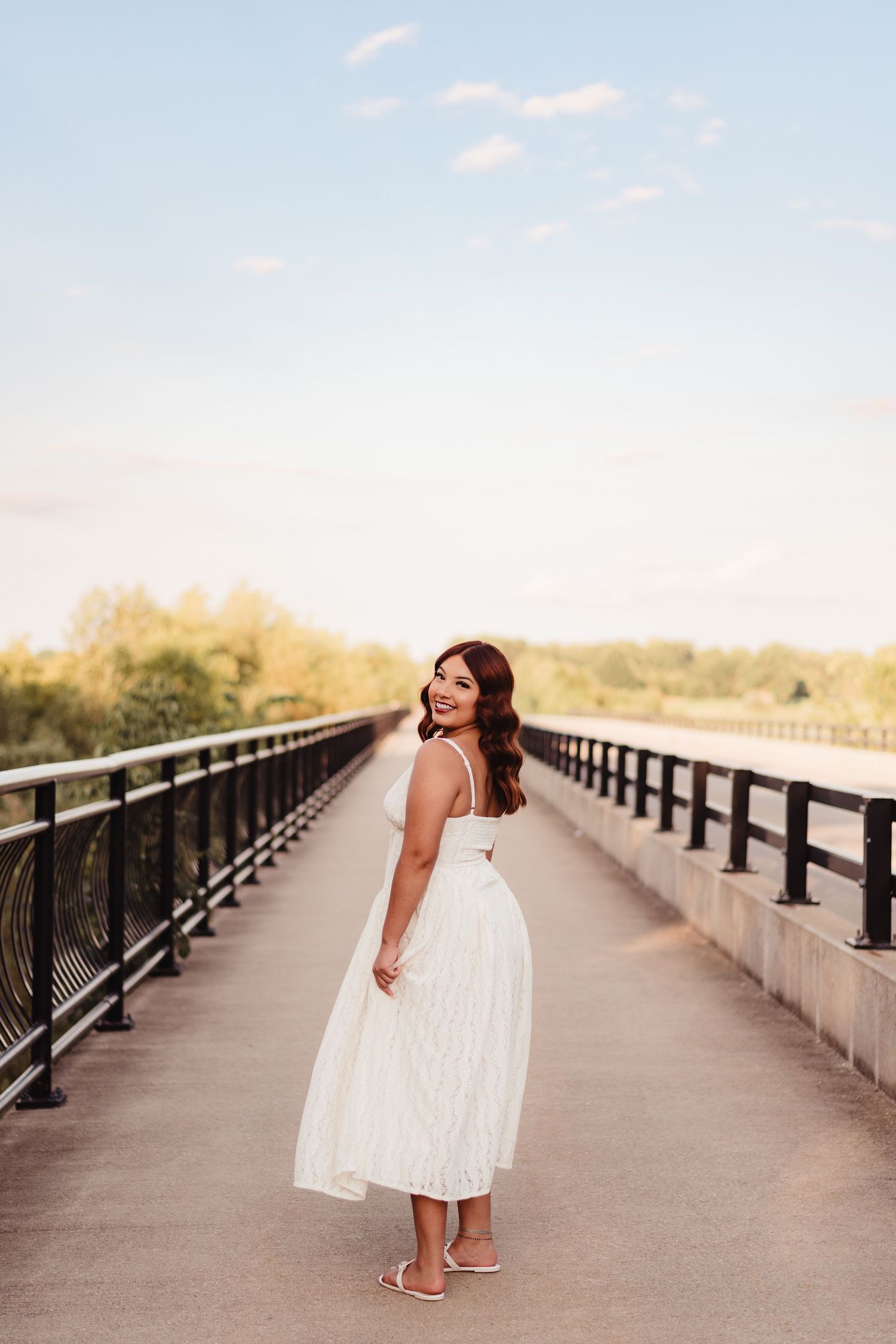 Woman in white midi dress and heels poses on a bridge at golden hour, smiling over her shoulder.