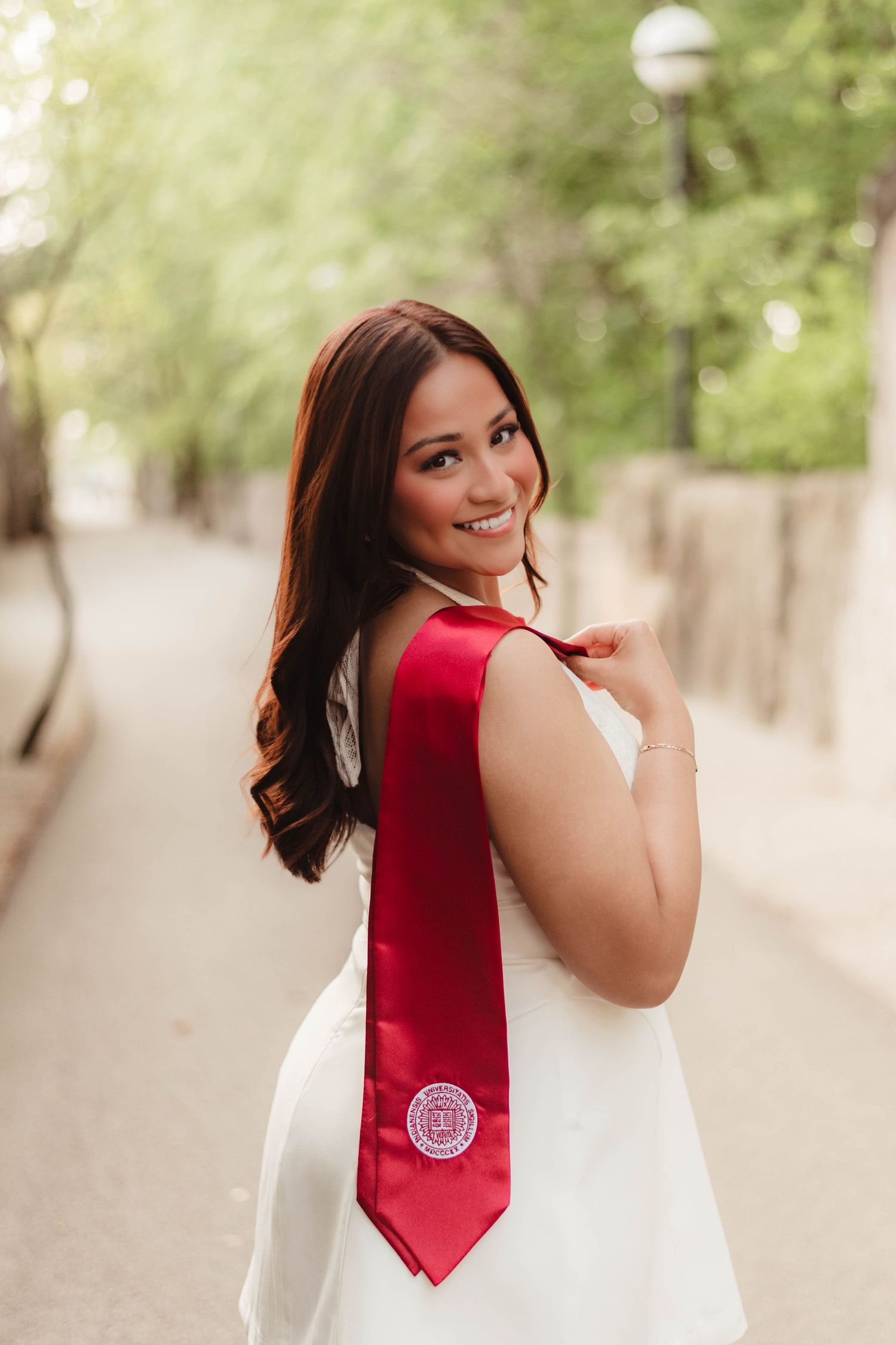 Smiling graduate poses outdoors in white dress with red university graduation sash draped over shoulder.