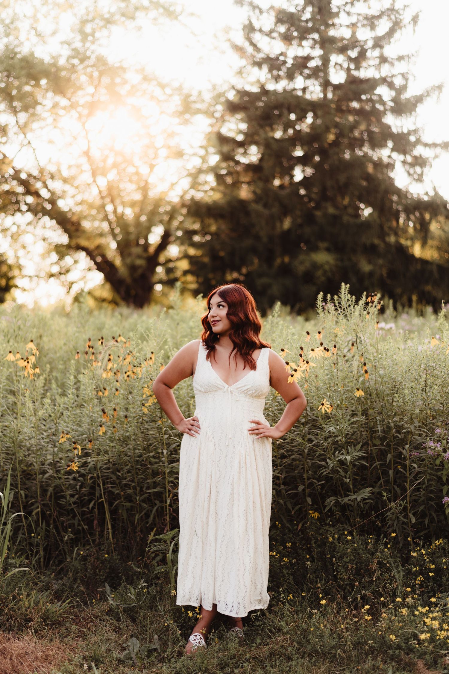 Woman in white midi dress standing in wildflower field at golden hour sunset with lush green trees background.
