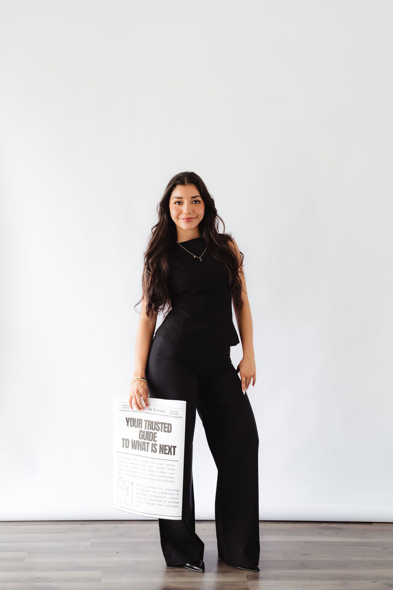 Woman in black outfit holding a newspaper with text 'Your Trusted Guide To What Is Next' against white wall.
