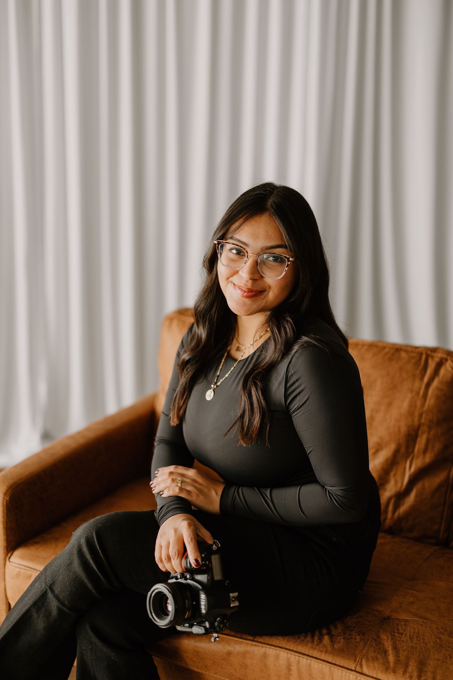 Woman in black outfit poses on brown leather couch for professional portraits in front of white curtains.