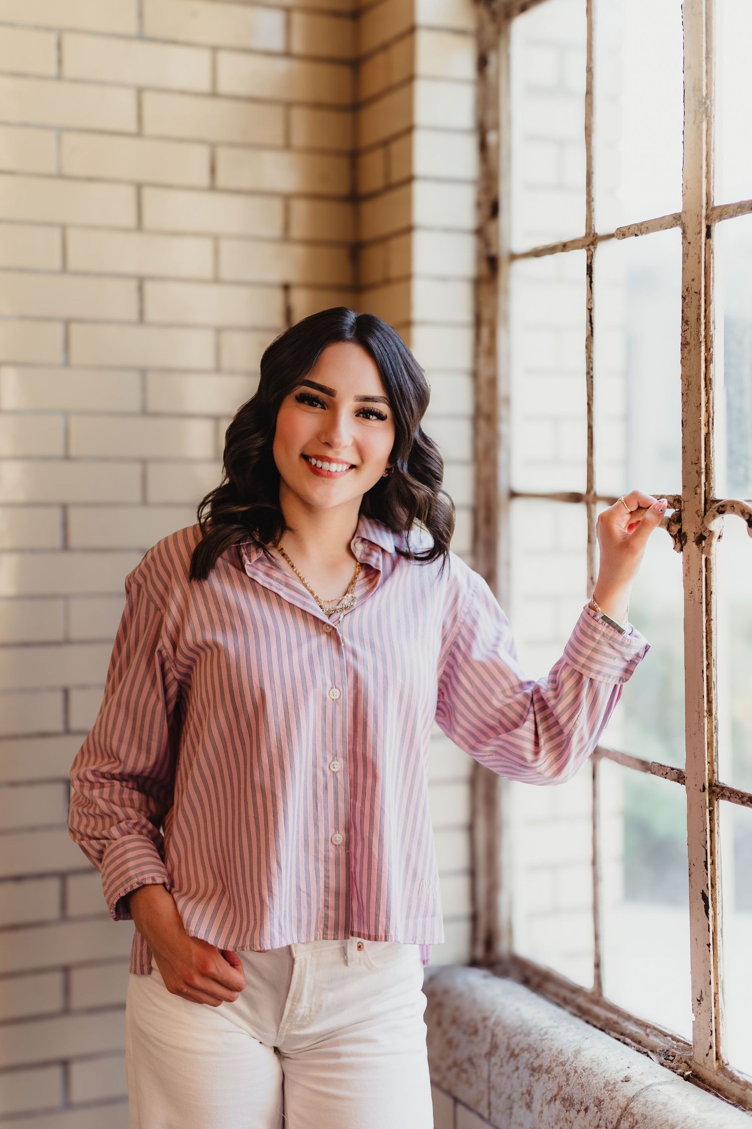 Smiling young woman in pink striped shirt and white jeans standing by a rustic window with brick wall background.