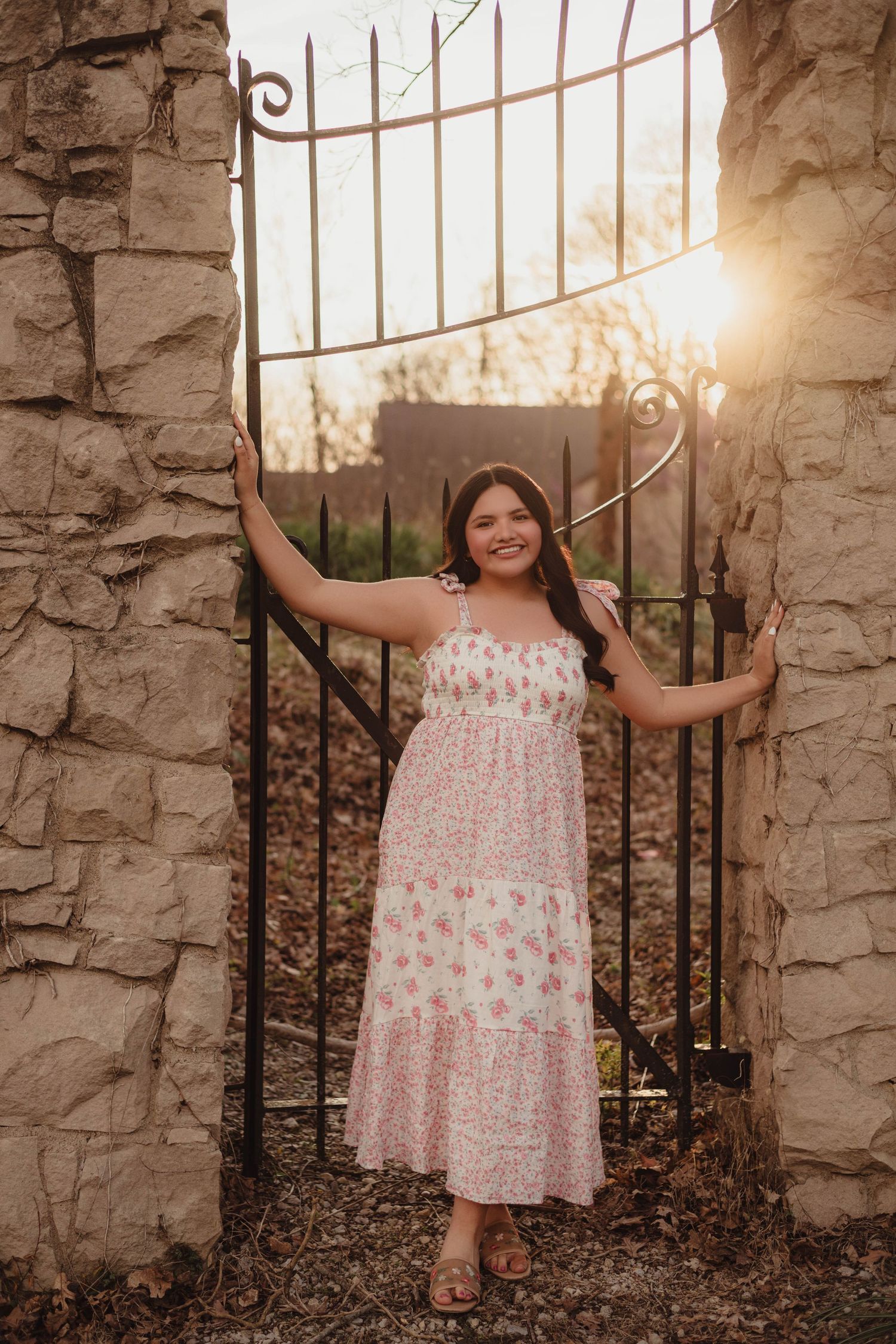 Senior in floral maxi dress posing at ornate iron gate between stone pillars at golden hour sunset in Lafayette, Indiana.