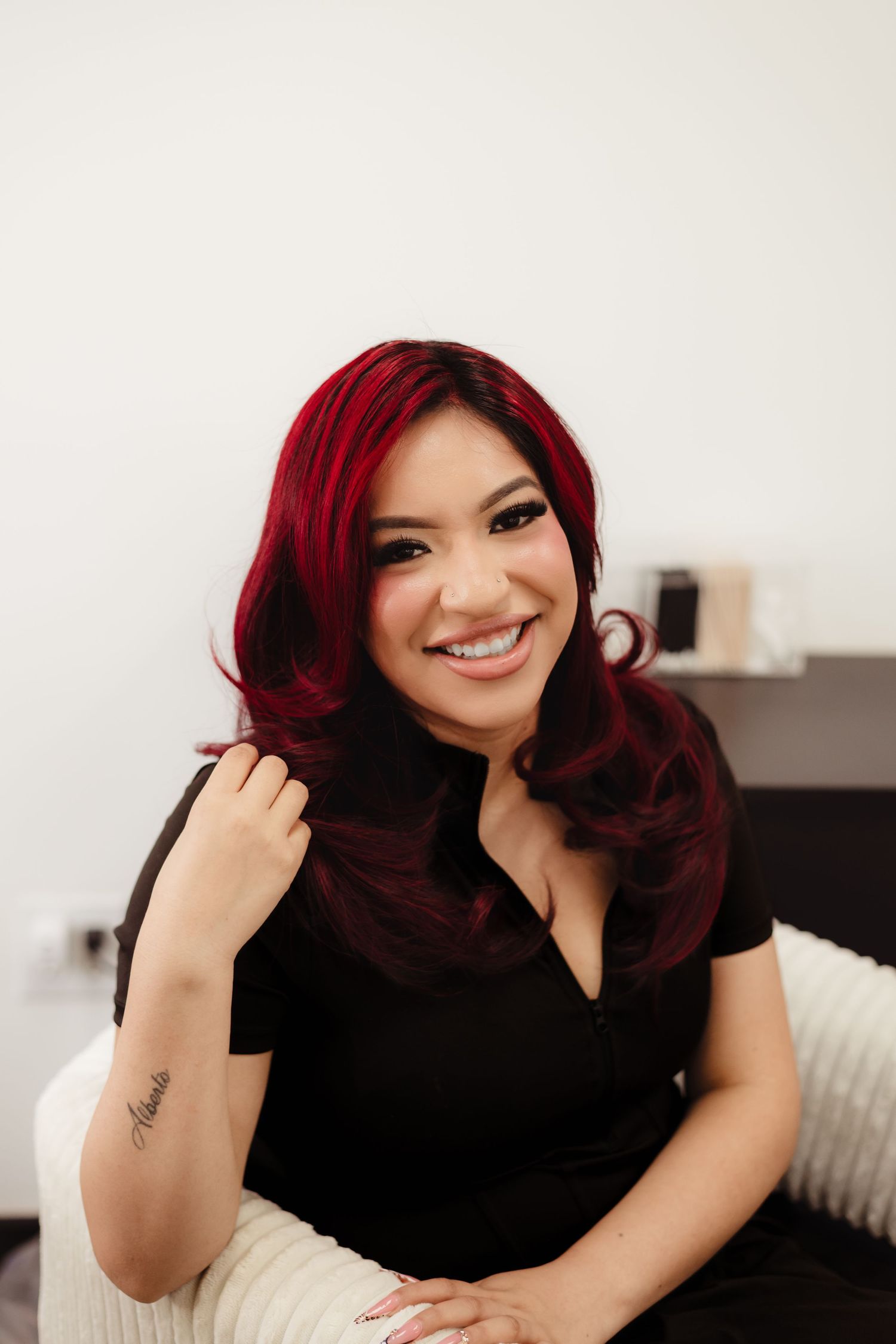 Smiling woman with vibrant red hair wearing a black top, sitting confidently in a bright modern room.