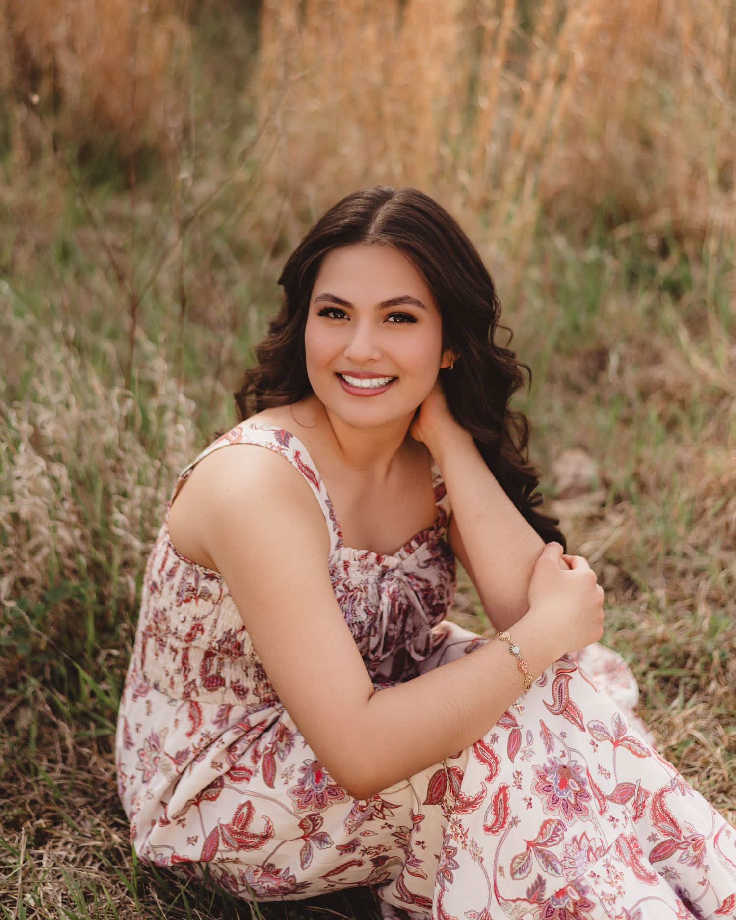 Young woman in floral dress sitting in golden grass field, smiling with hand on cheek in outdoor portrait in Lafayette, indiana.