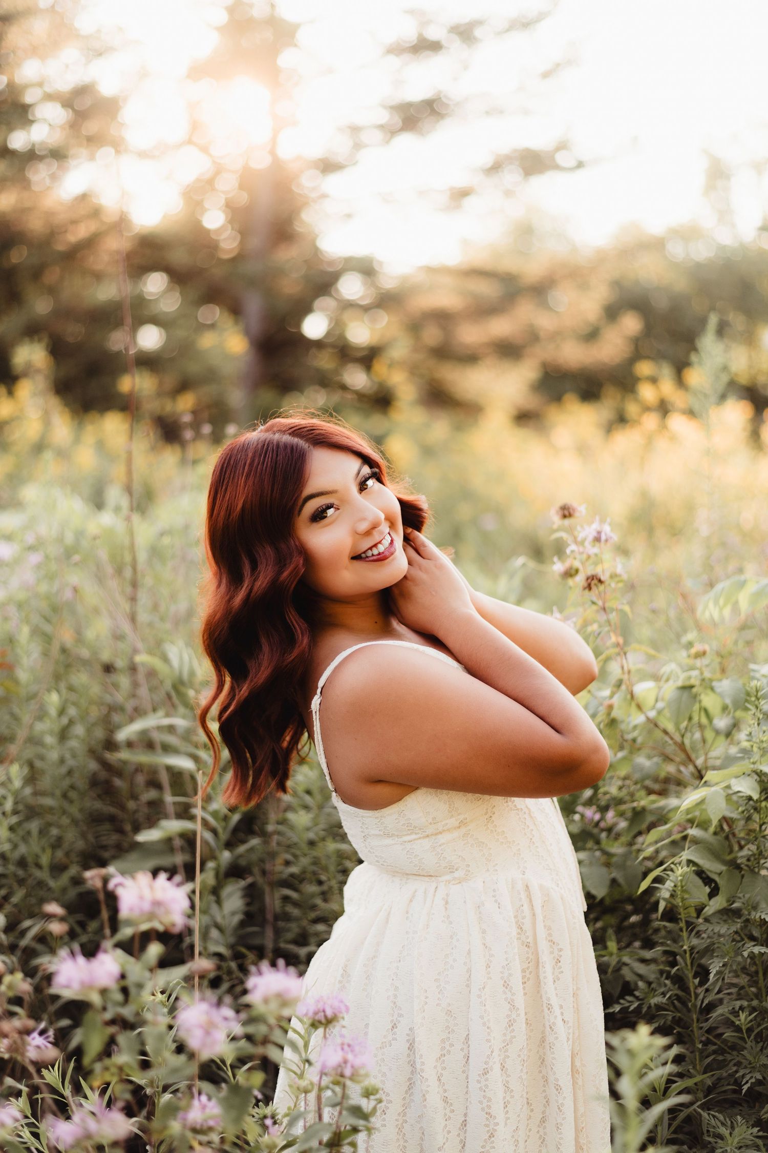 Young woman in white lace dress smiling in a wildflower field at golden hour sunset.