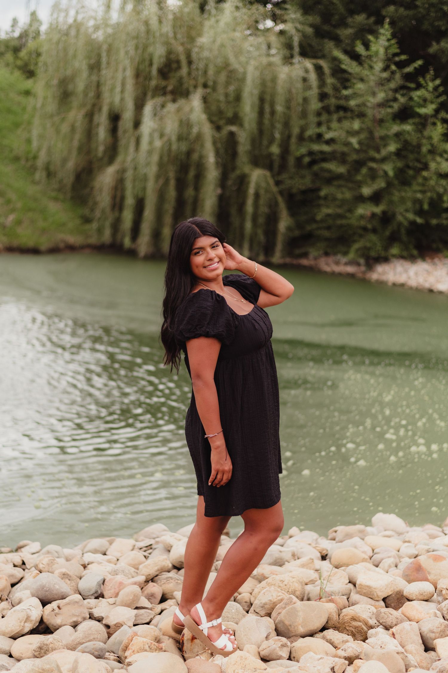 A person poses joyfully on rocky terrain beside a serene green lake surrounded by willow trees.