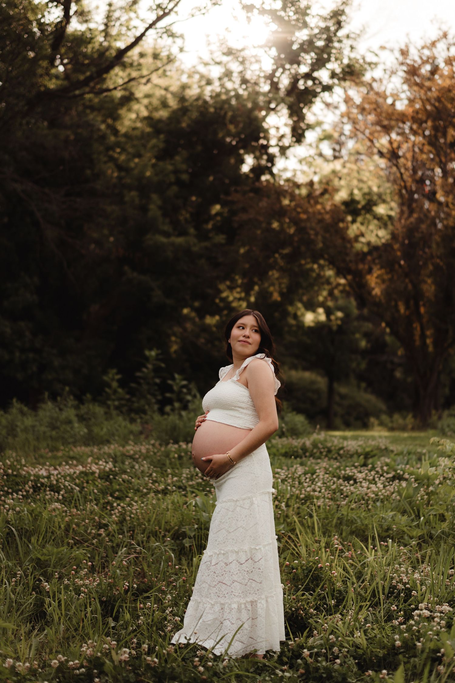 Pregnant woman in white lace dress standing in field at golden hour.