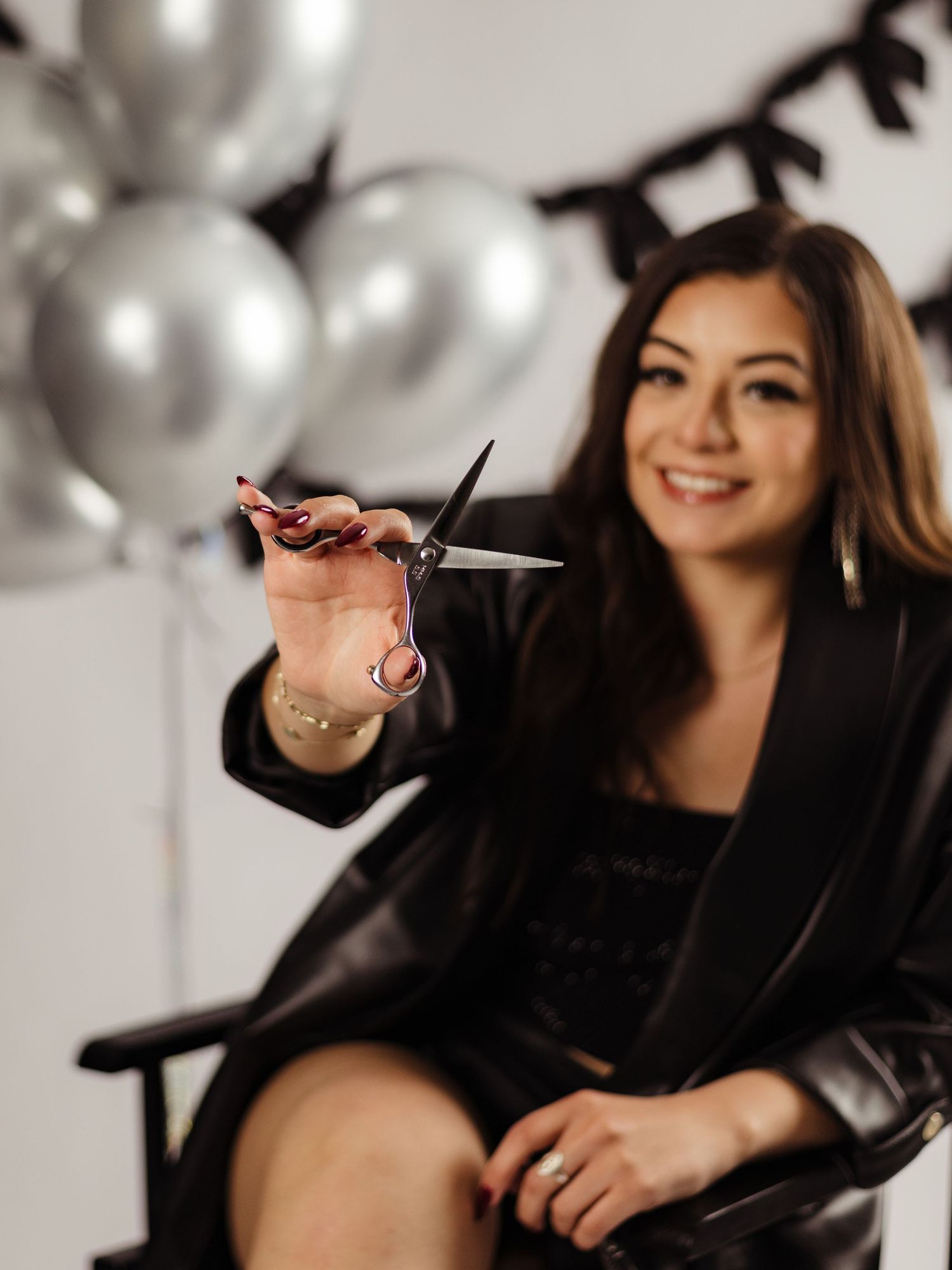 Smiling female hairstylist holding scissors toward camera, seated in chair with silver balloons in background.