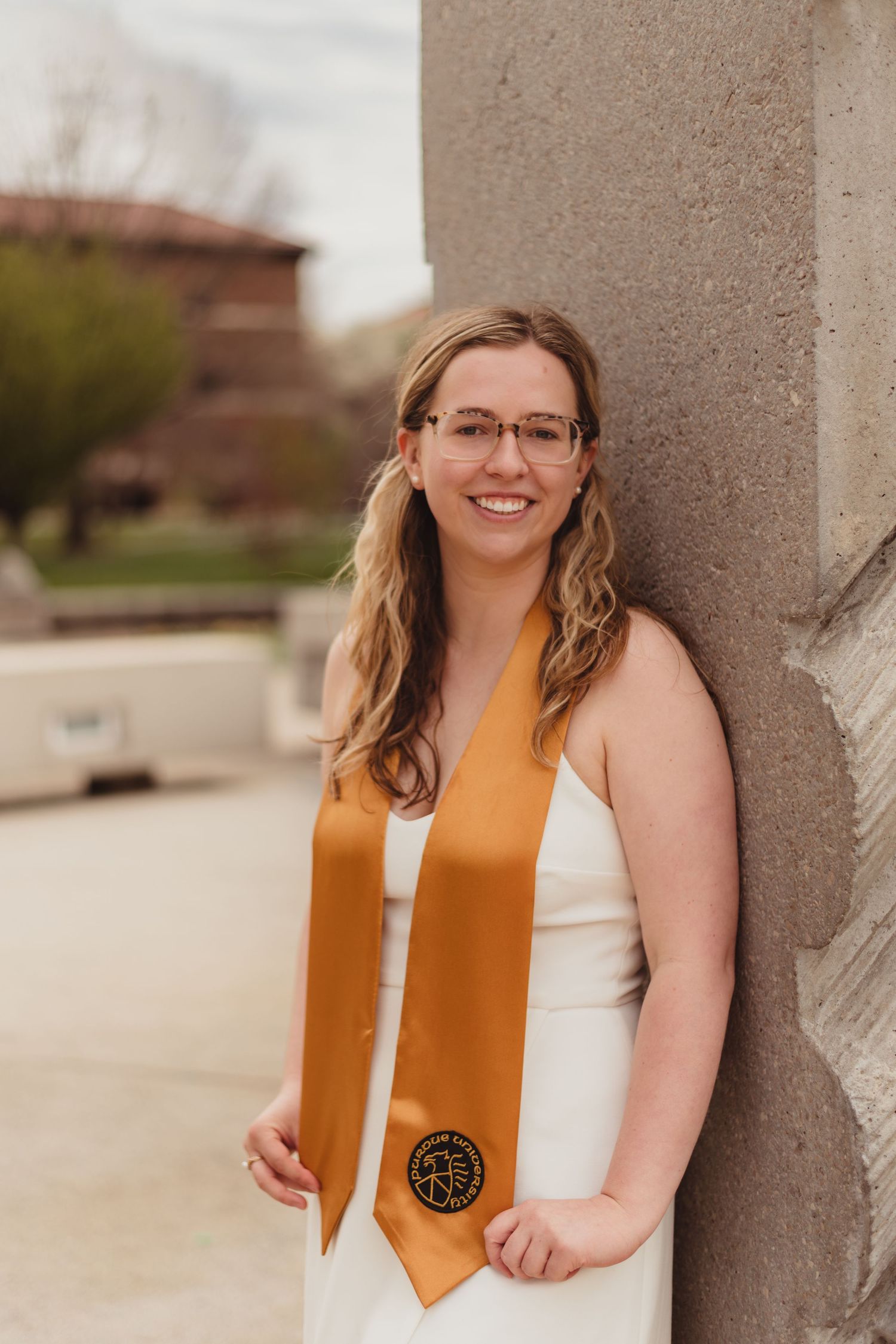 Graduate in white dress and orange graduation stole stands smiling against stone column at Purdue Campus in West Lafayette, IN