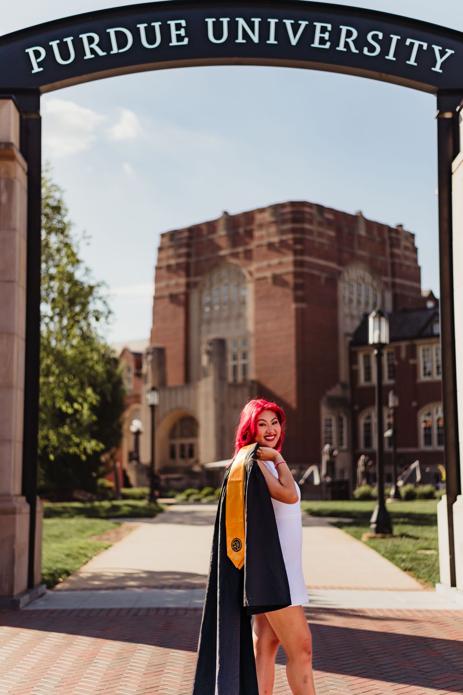 Graduate poses in cap and gown in front of Purdue University buildings on a sunny day.