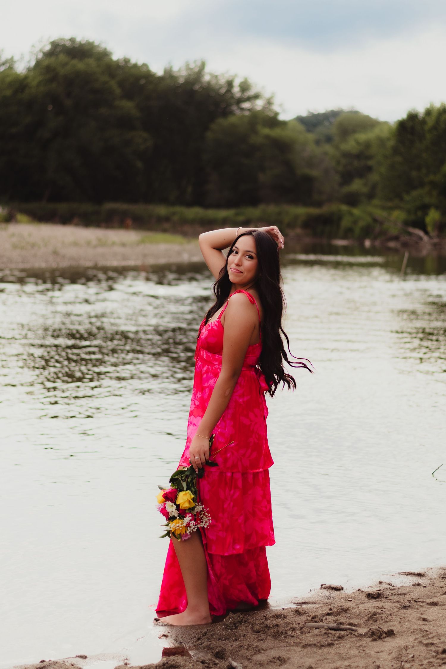 A woman in a vibrant red lace dress poses gracefully by a serene lakeside with a colorful bouquet of flowers.