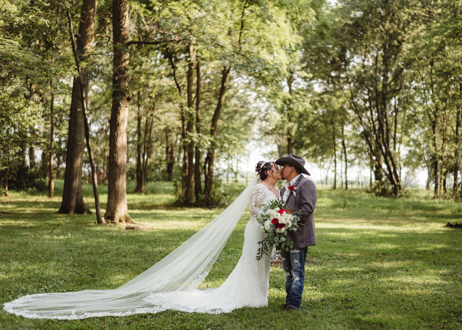 A couple in wedding attire share an intimate moment in a sunlit wooded area with a long flowing veil in Lafayette, Indiana.