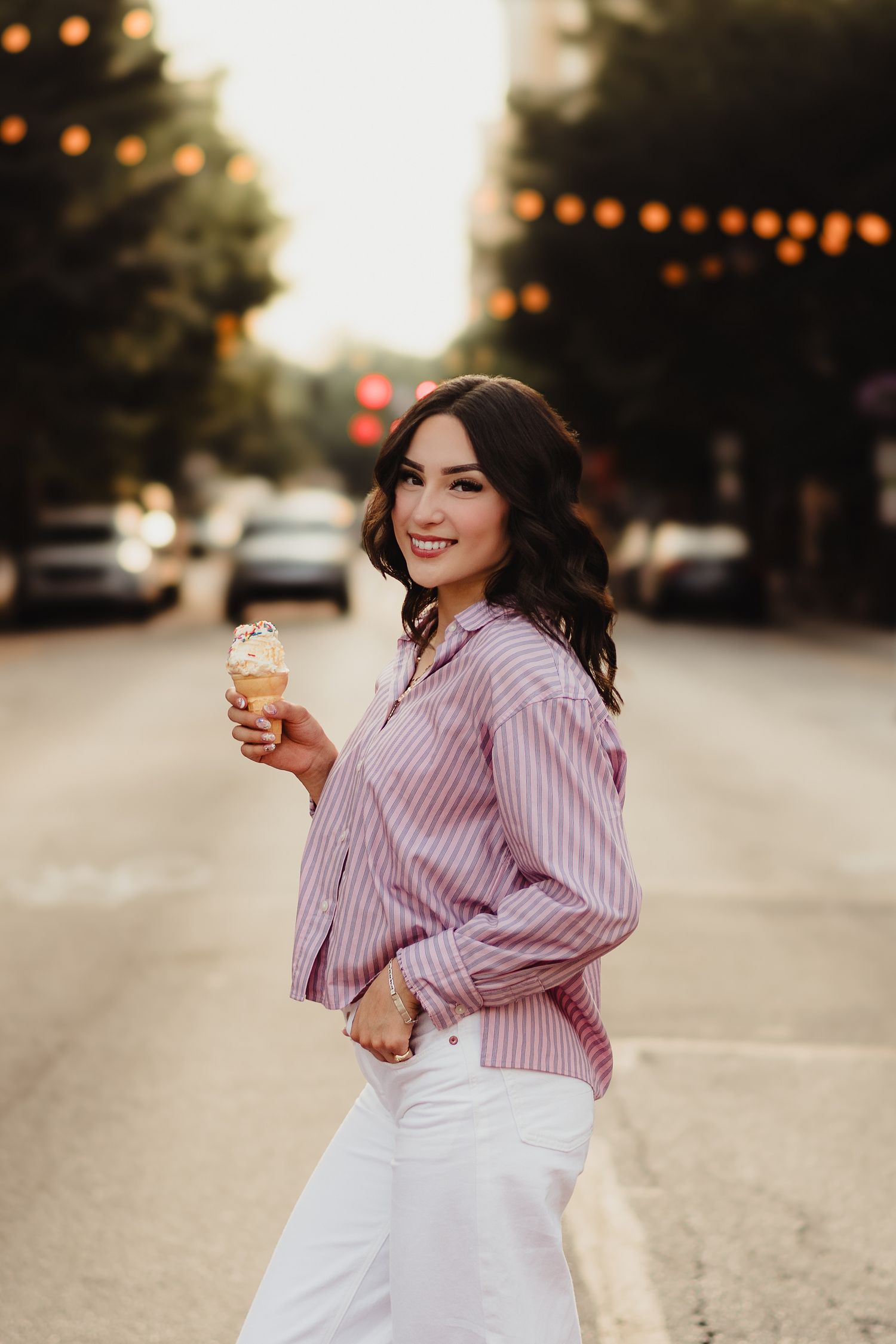 Woman in pink striped shirt and white jeans holding ice cream cone on a street with string lights.