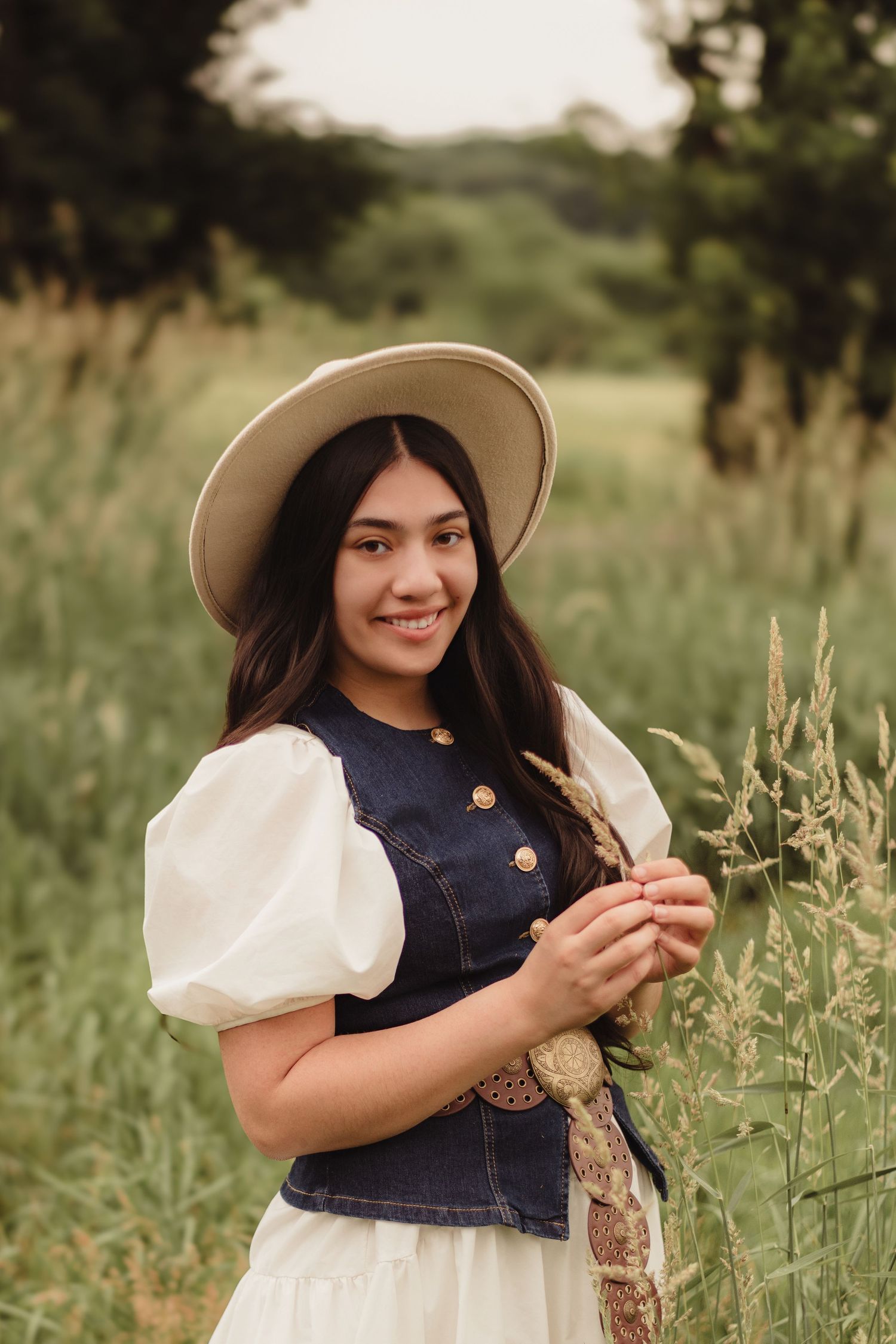 Smiling teen girl in denim vest, white dress, and wide-brim hat poses in tall grass field.
