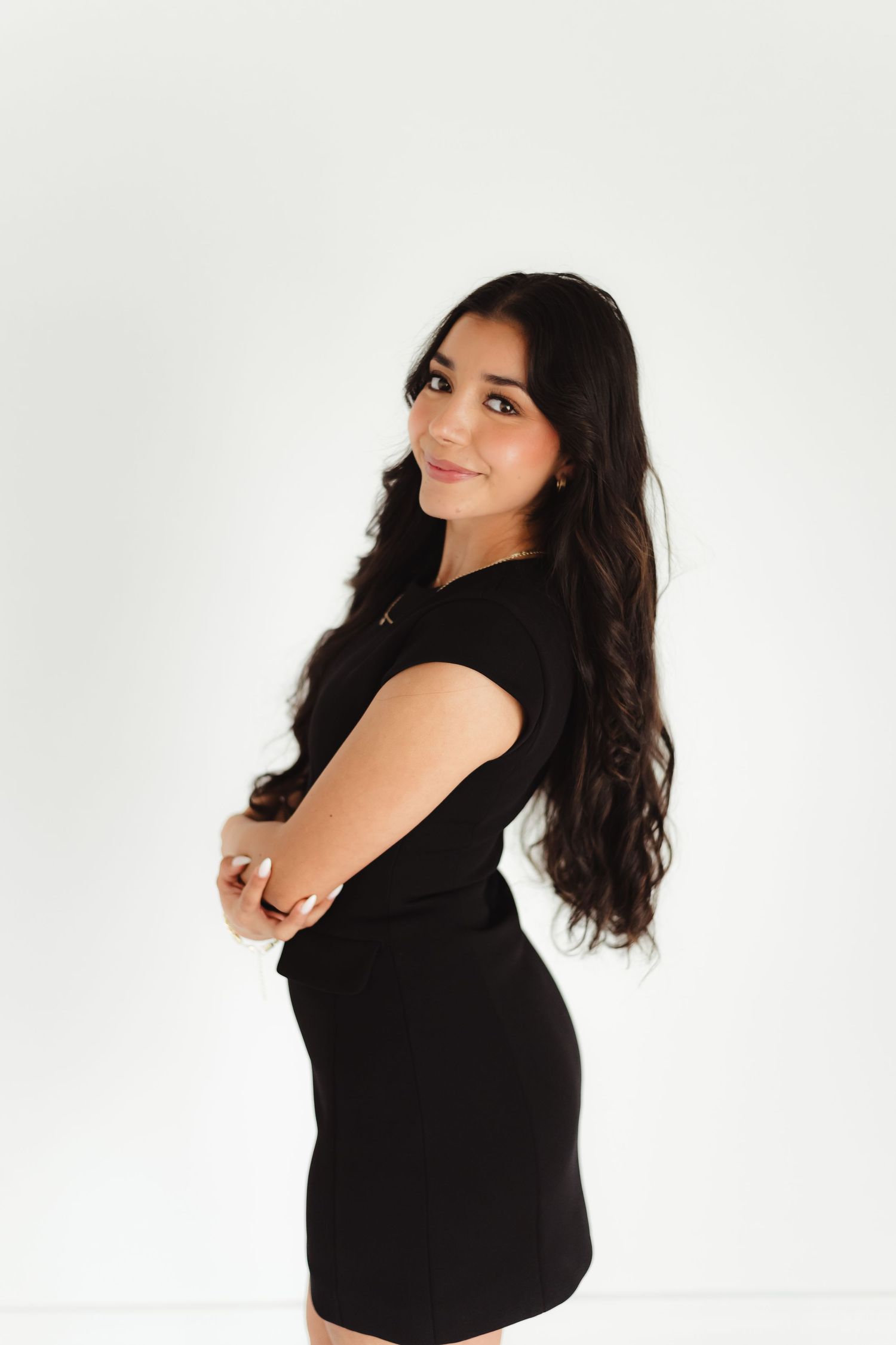 Young woman with long dark wavy hair poses in a black mini dress against a white background.