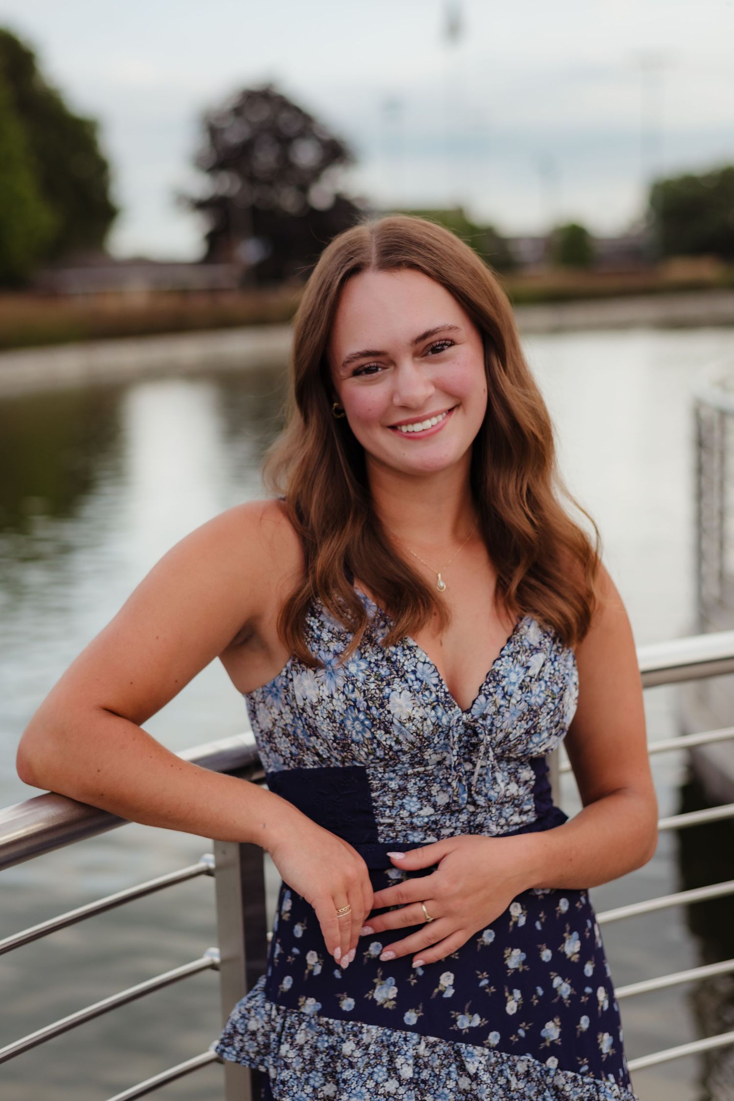 A person in a navy blue and white floral dress poses along a waterfront railing during a photo session.