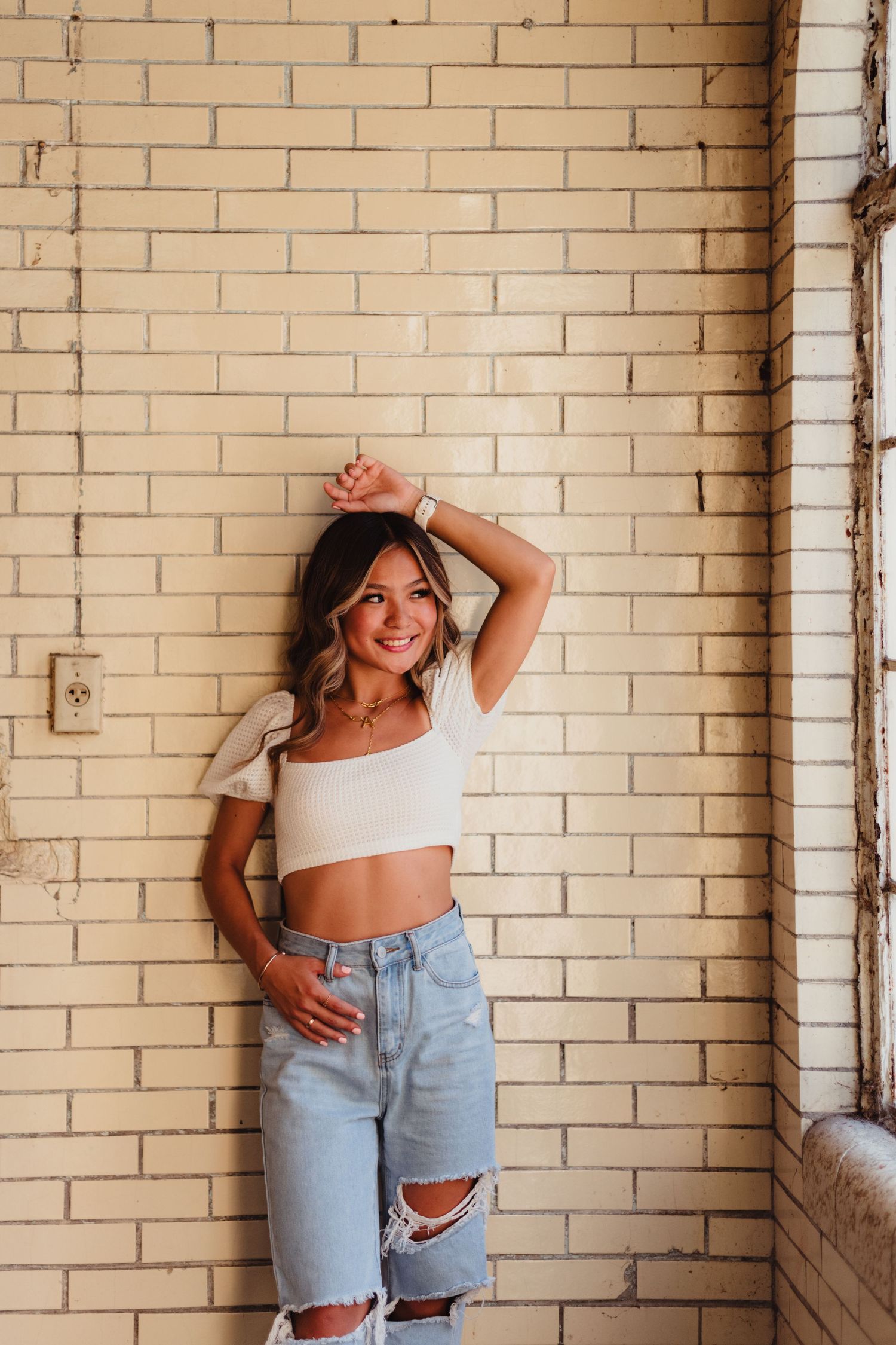 Woman in white crop top and ripped denim shorts poses against brick wall in casual photoshoot.