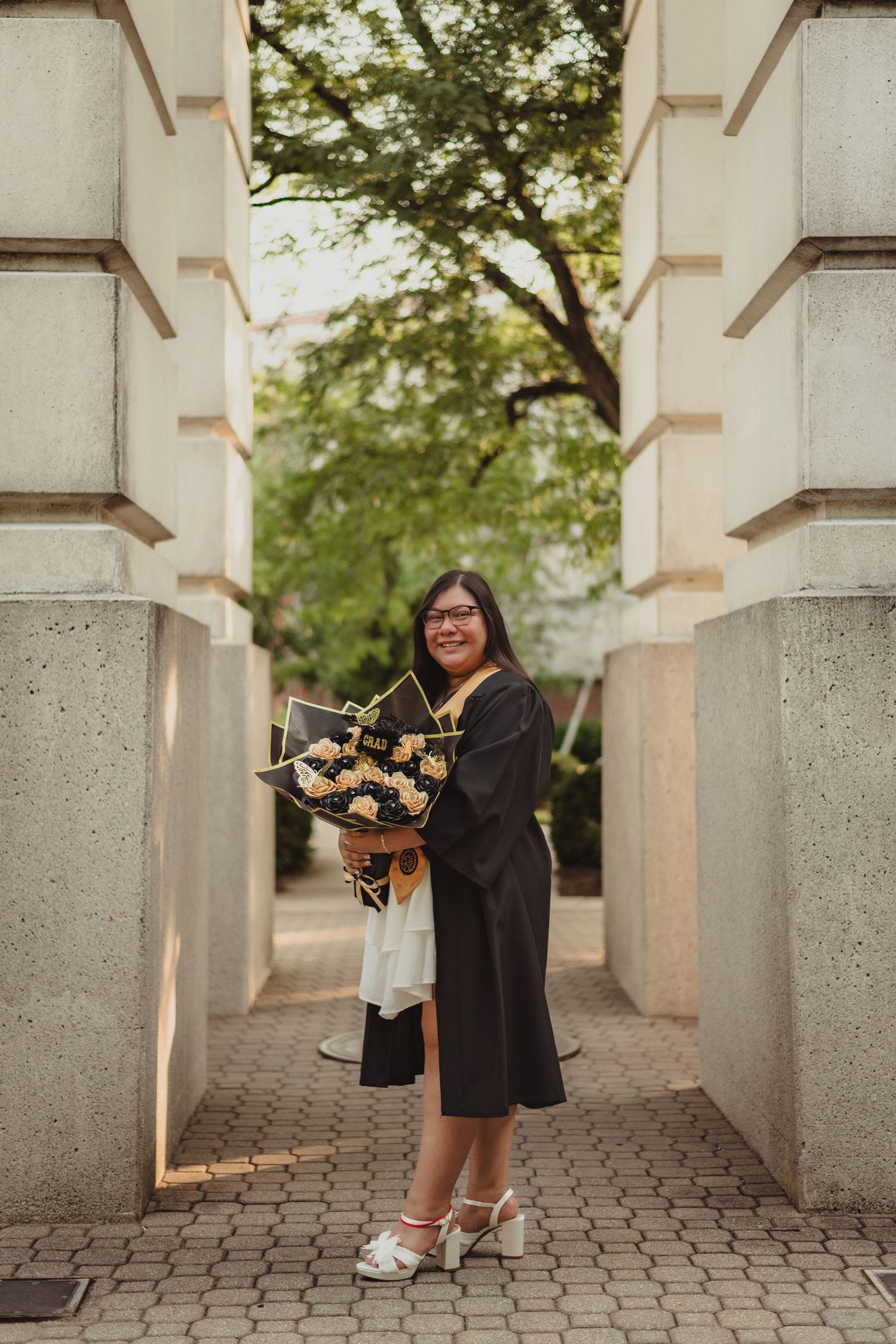 Graduate poses playfully between concrete columns wearing black dress and holding graduation cap in outdoor setting  at Purdue Campus.