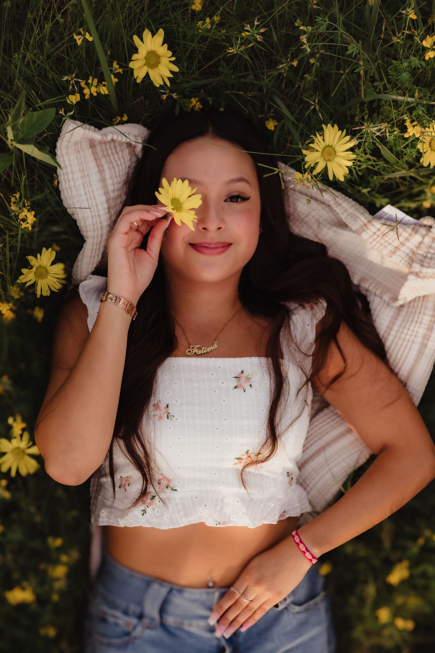 A lifestyle portrait of someone lying in a field of yellow daisies holding a flower to their face.