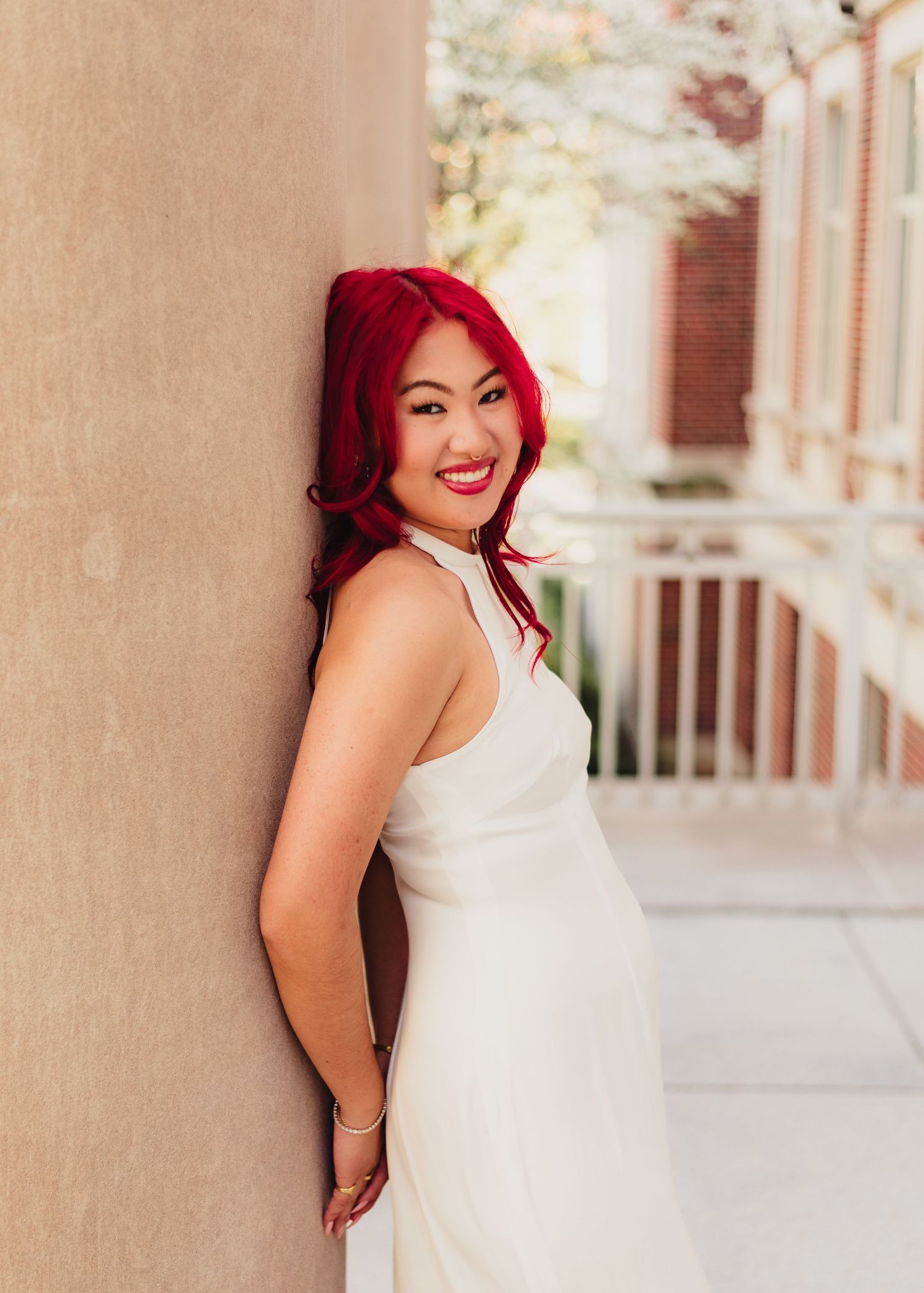 A person with bright red hair wearing a white dress poses against a beige stone column in an elegant setting at Purdue Campus in West Lafay