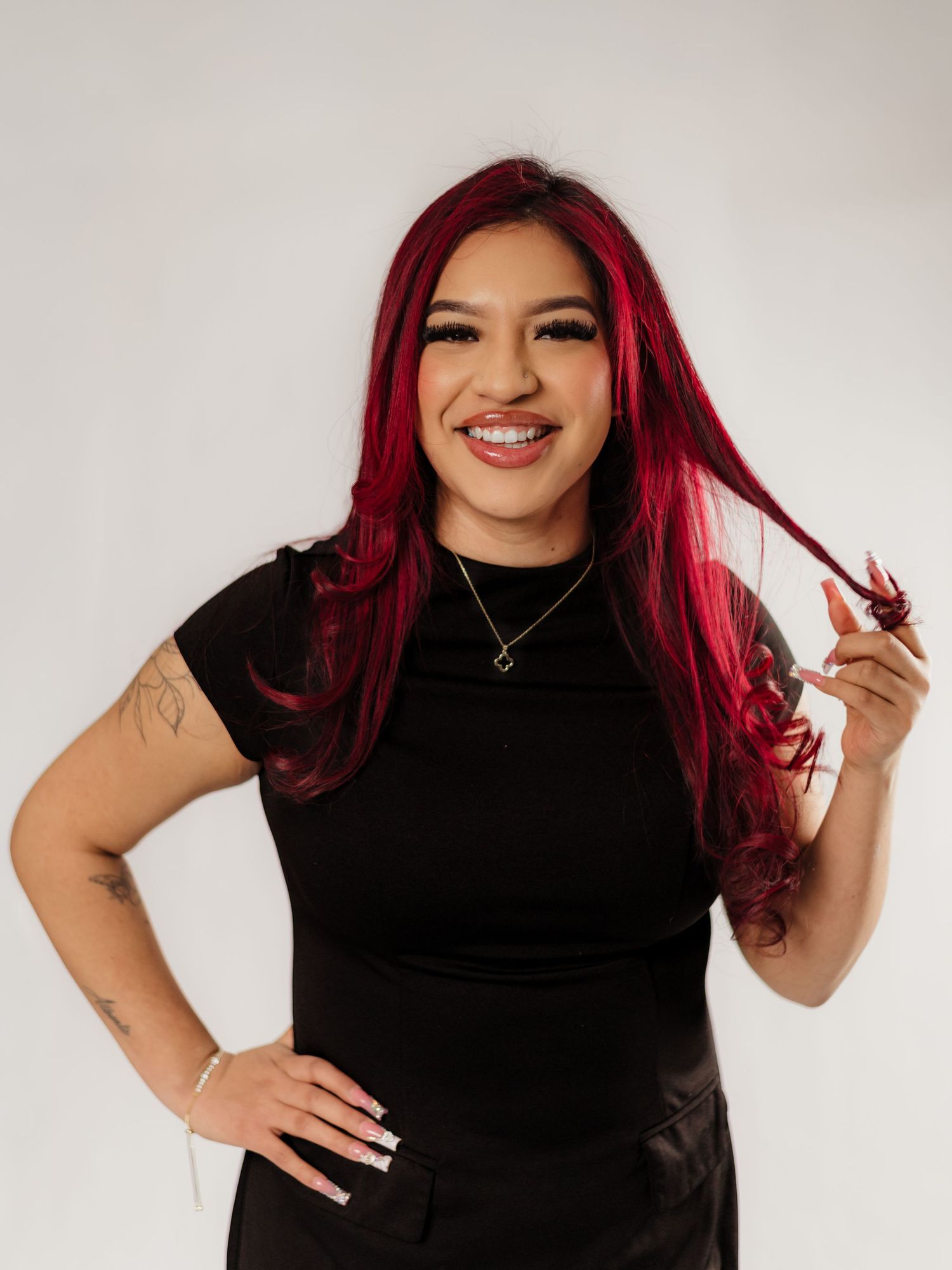 Smiling woman with vibrant red hair in a black outfit, holding her curled hair ends, with a tattoo on her arm.