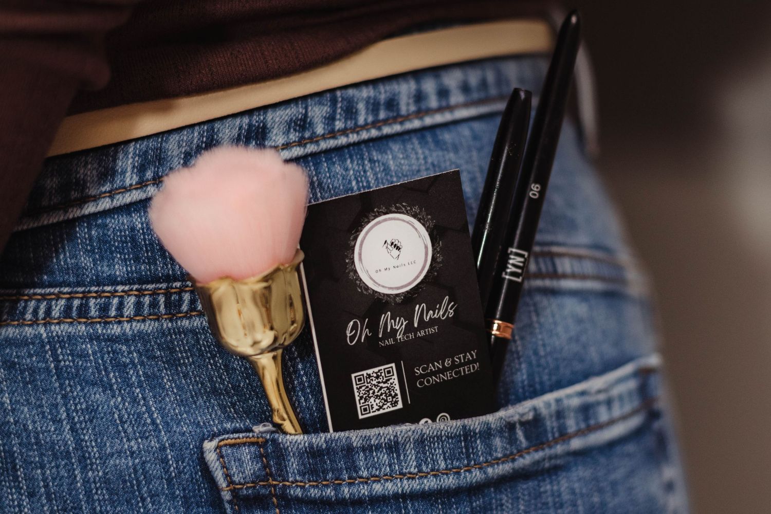 Pink makeup brush and Oh My Nails business card with QR code tucked into denim jeans back pocket.