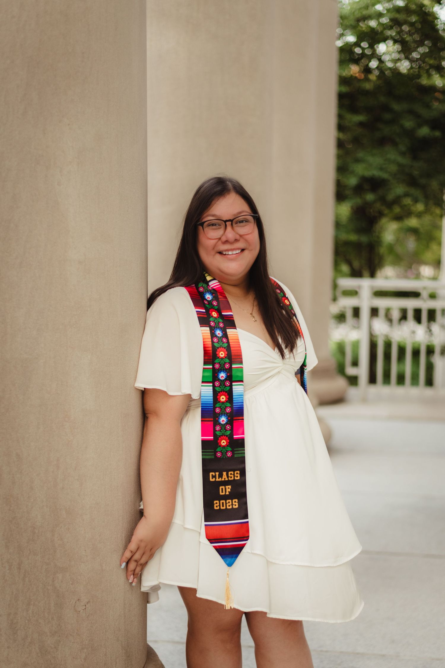 Graduate in white outfit poses with decorated graduation stole and colorful stickers against stone columns  at Purdue Campus.