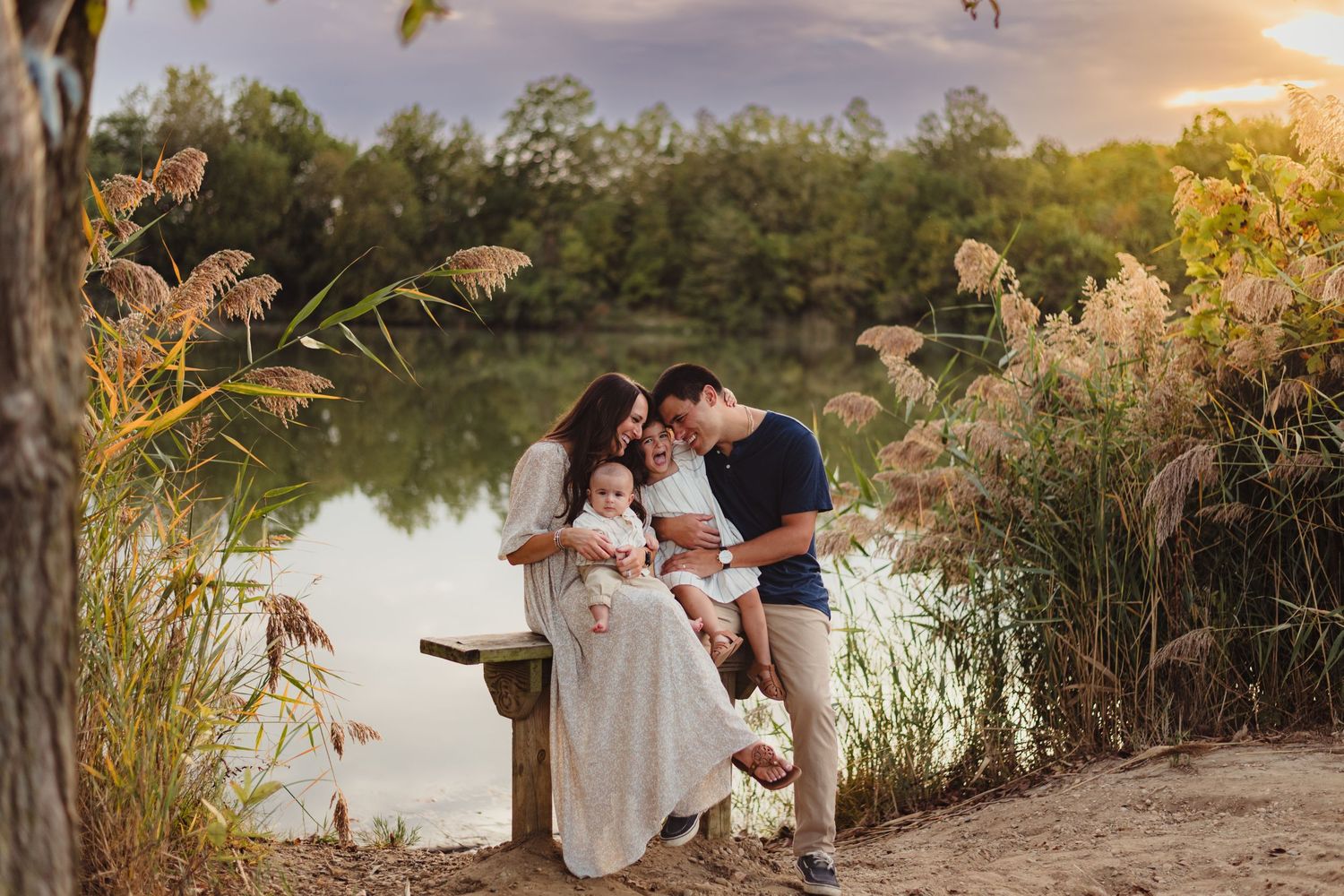Family with two babies shares tender moment on bench beside serene lake at golden sunset.