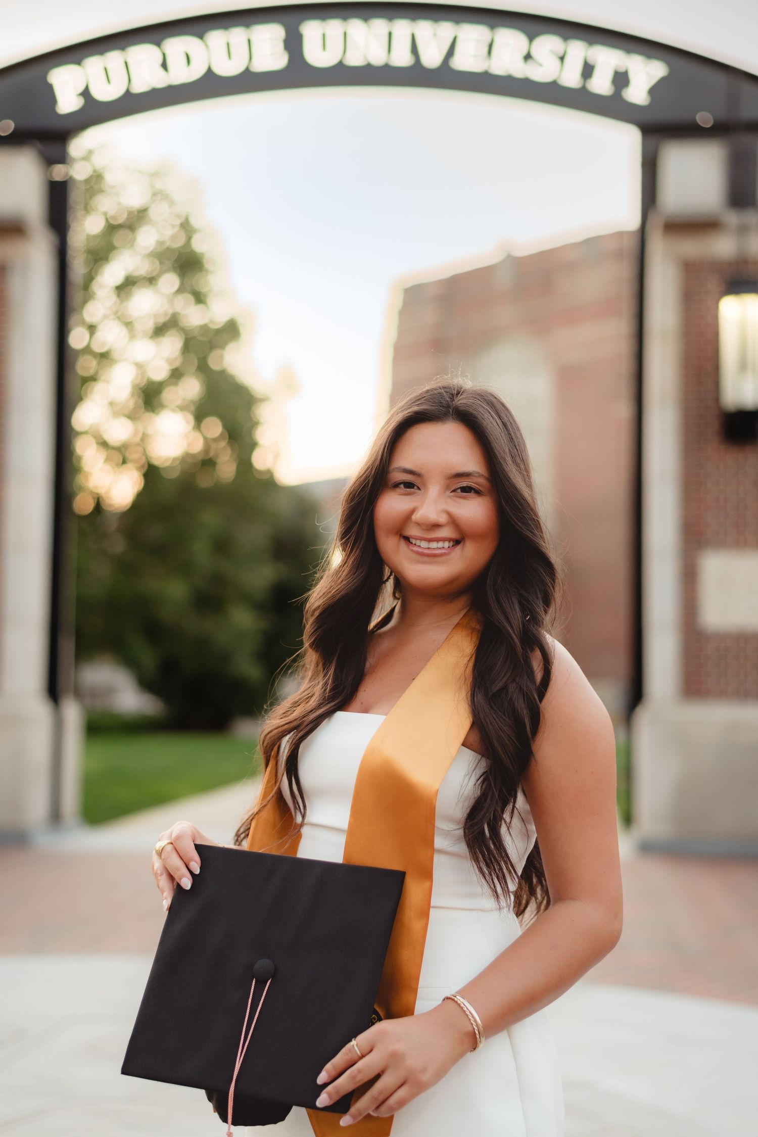 A graduate in an orange top and black gown poses with their diploma on a university campus during sunset  at Purdue Campus in West Lafayette