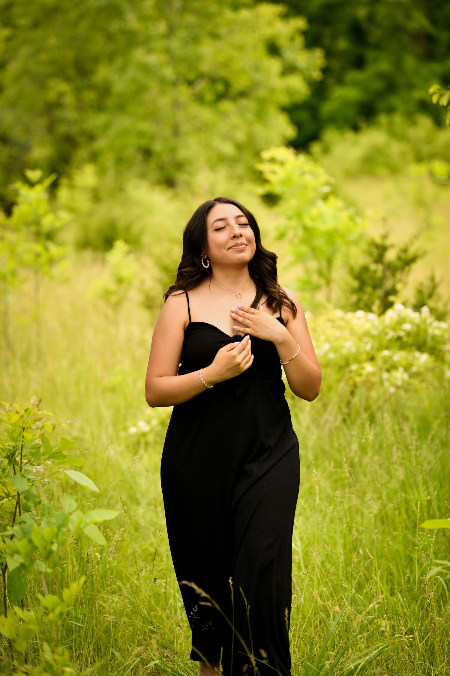 A person in a black dress poses expressively in a sunny meadow with green foliage and wildflowers in the background.