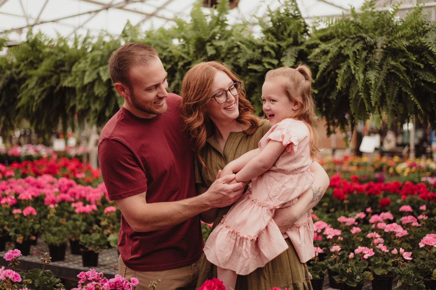 A family shares laughter together in a greenhouse filled with pink flowers and hanging plants.