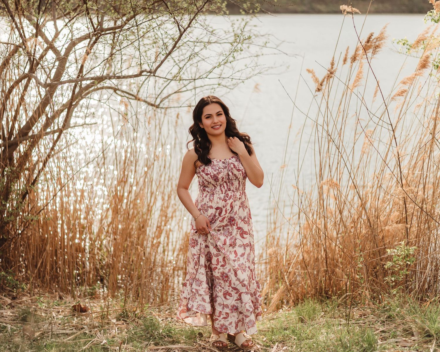 High school Senior in floral maxi dress standing by a sunlit lake surrounded by tall grasses and bare trees in Lafayette, indiana