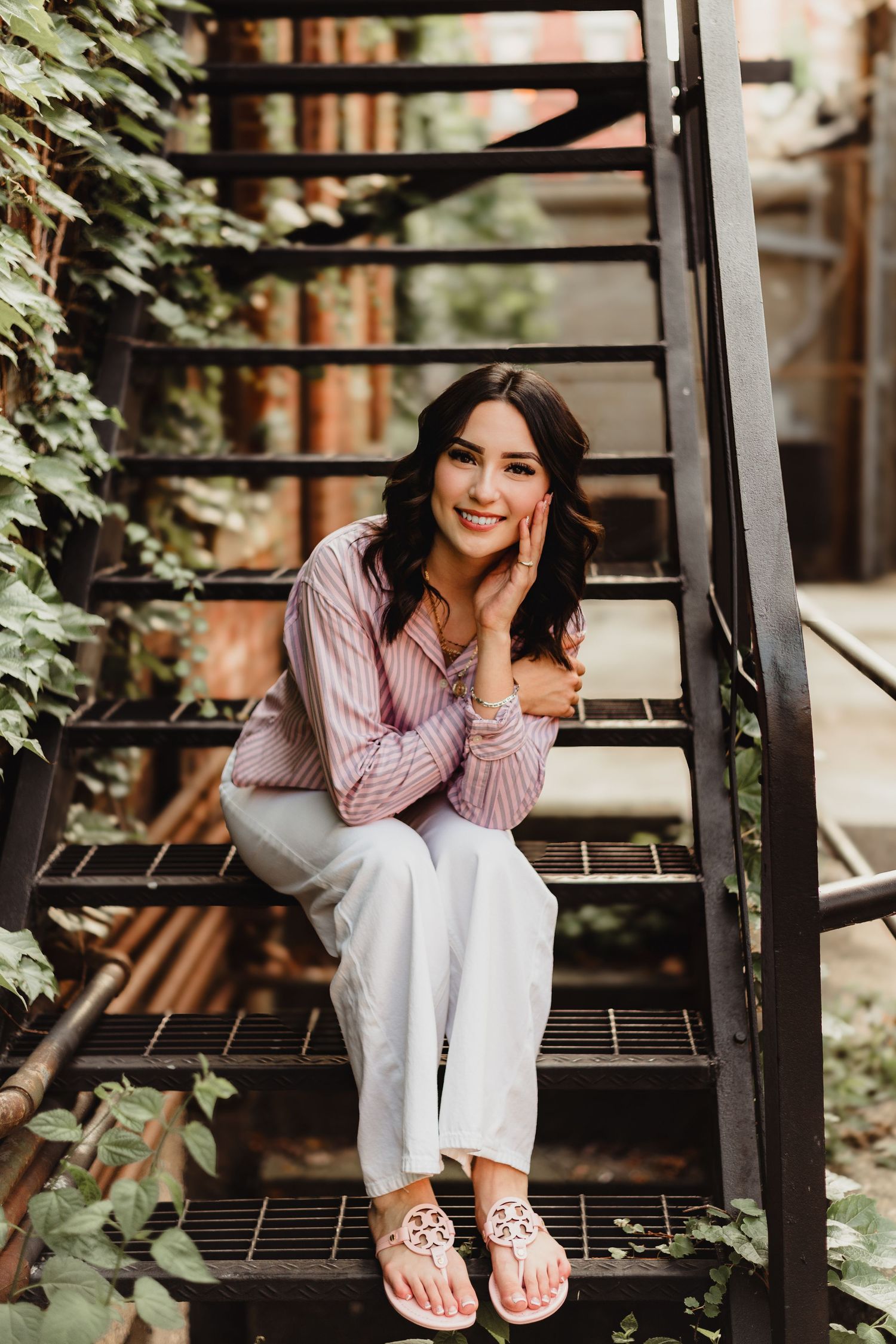 Smiling brunette woman in pink striped shirt and white pants sitting on metal stairs surrounded by green ivy.