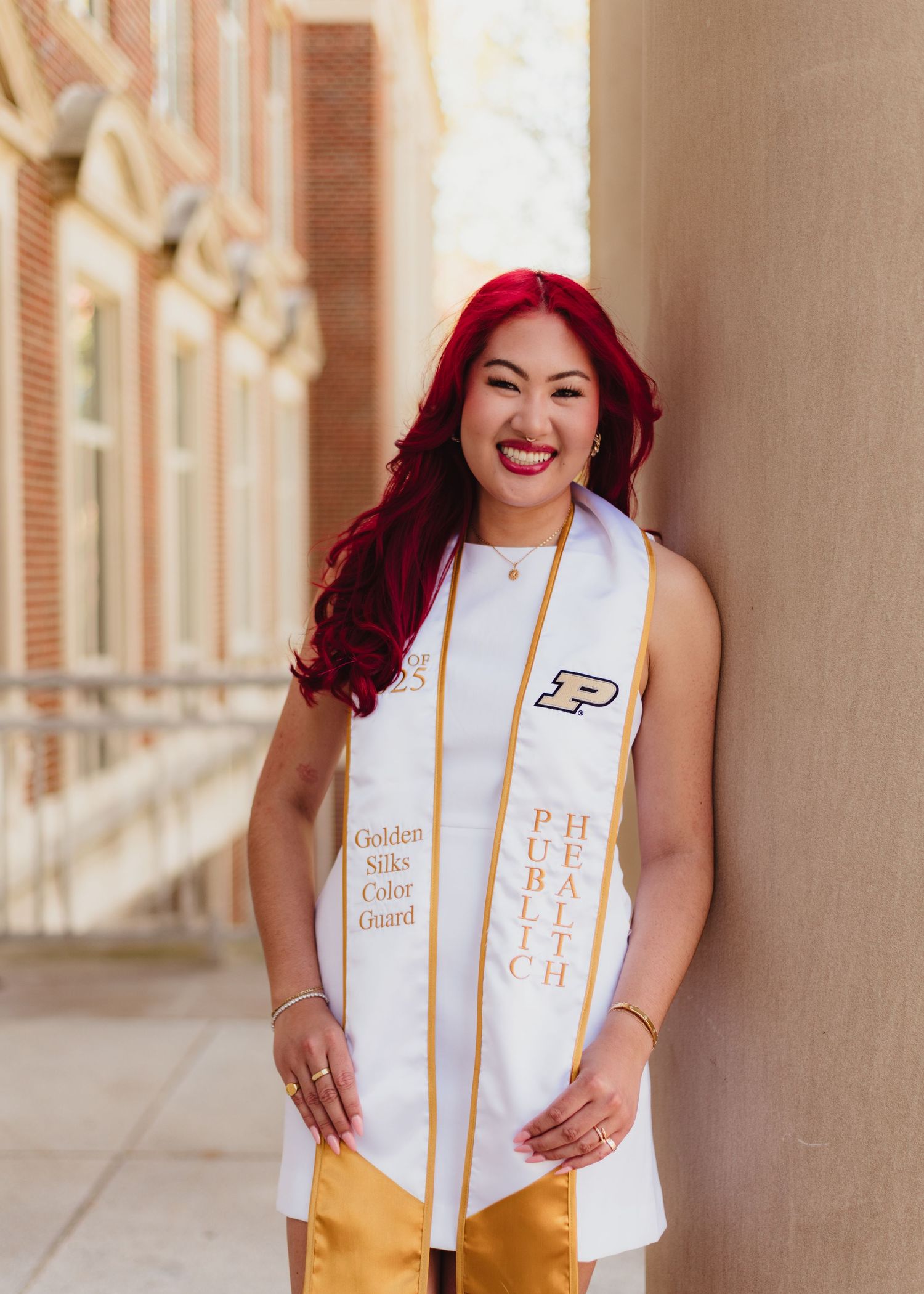 Graduate wearing white dress with gold stoles and red hair poses for graduation photos.