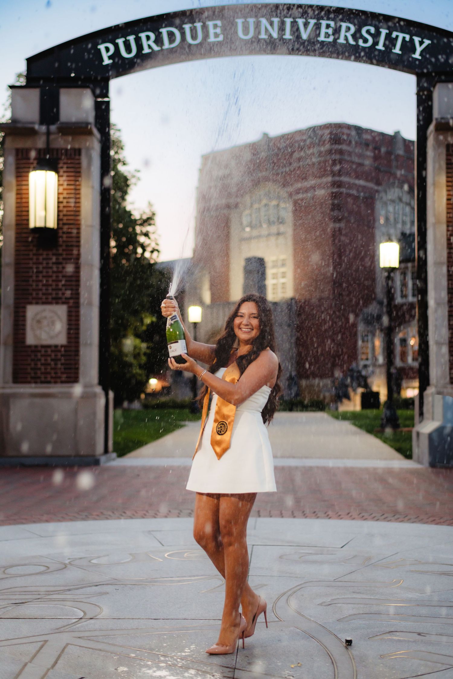 A graduate poses at Purdue University's entrance gates at dusk during a celebratory photo session.
