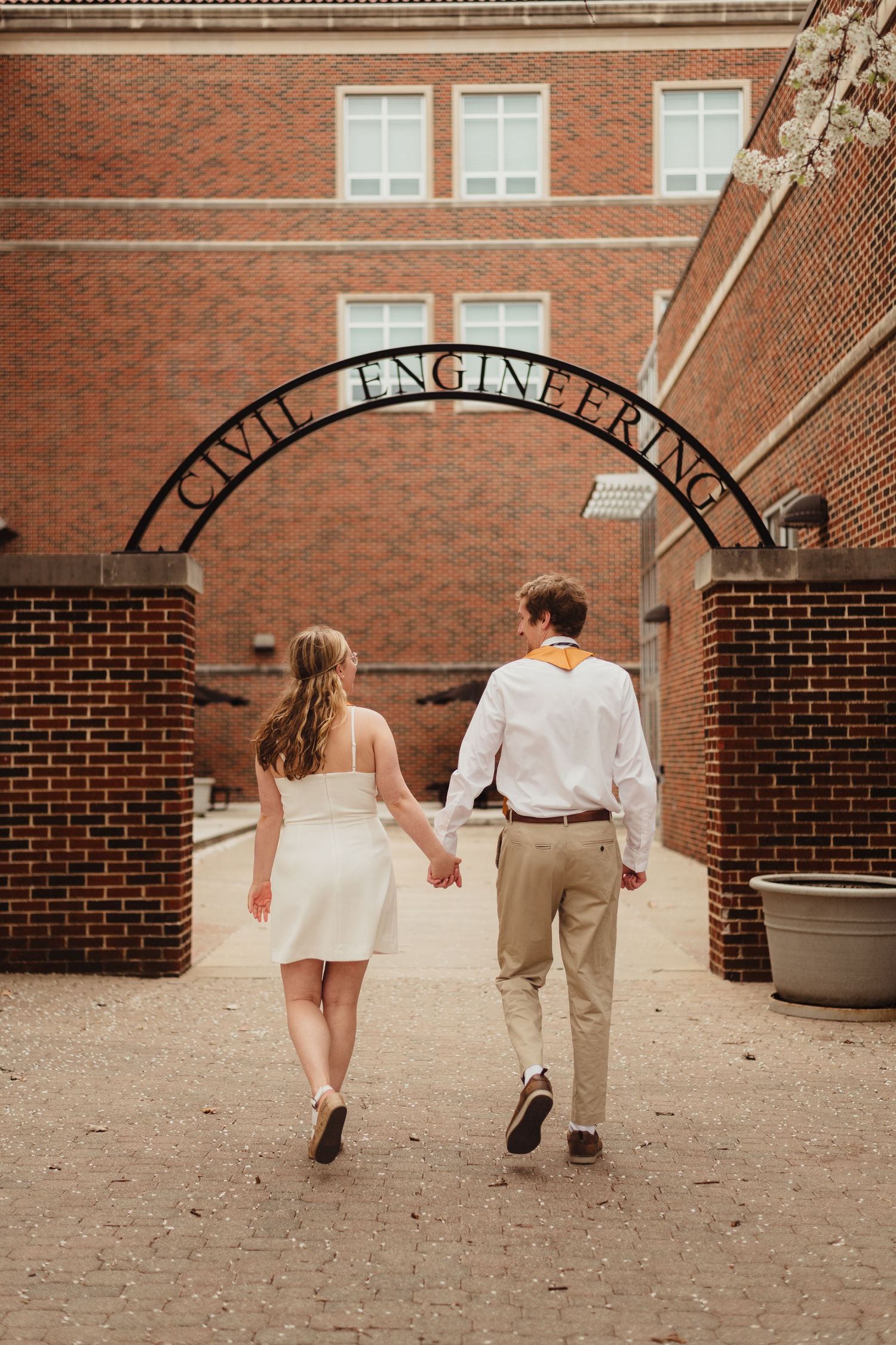 Couple walks hand in hand under brick archway on Purdue college campus during graduation celebration.