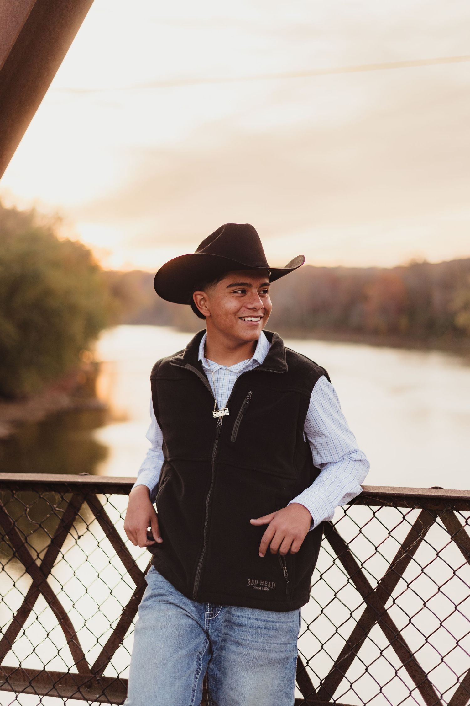 A person in a black vest and cowboy hat smiles while sitting on a fence during golden hour sunset.