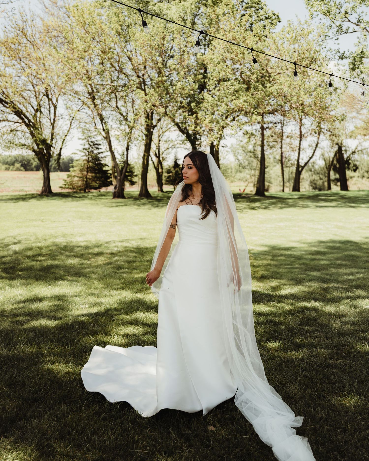 Bride in elegant white gown with long veil standing on lush green lawn surrounded by trees at outdoor wedding venue in Lafayette, Indiana.