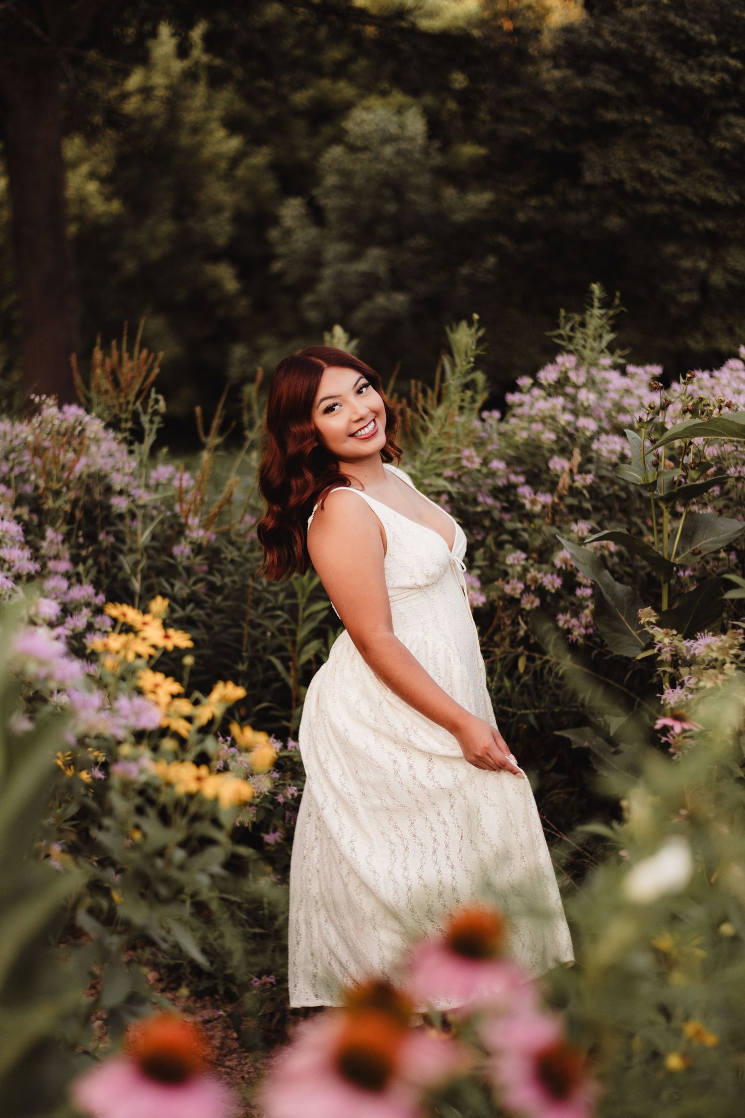 Young woman in white lace dress smiling among colorful wildflowers in a lush garden setting.